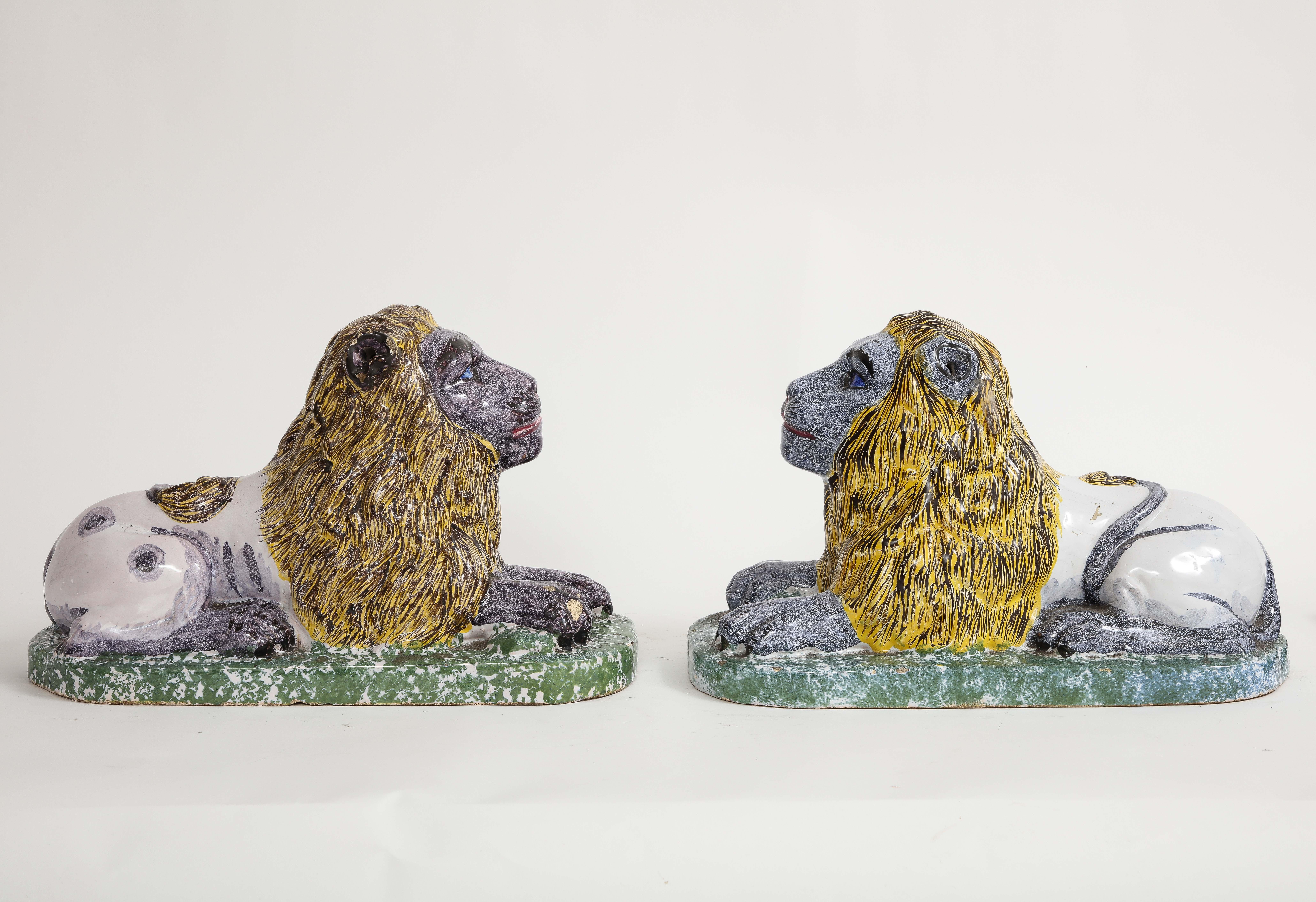 A Pair of 19th century French Majolica/Fiance Models of Lions Perched on stands. Each is wonderfully hand painted in vibrant purple and blue colorways. The bases are green and each is naturalistically looking forward perched on a rectangular