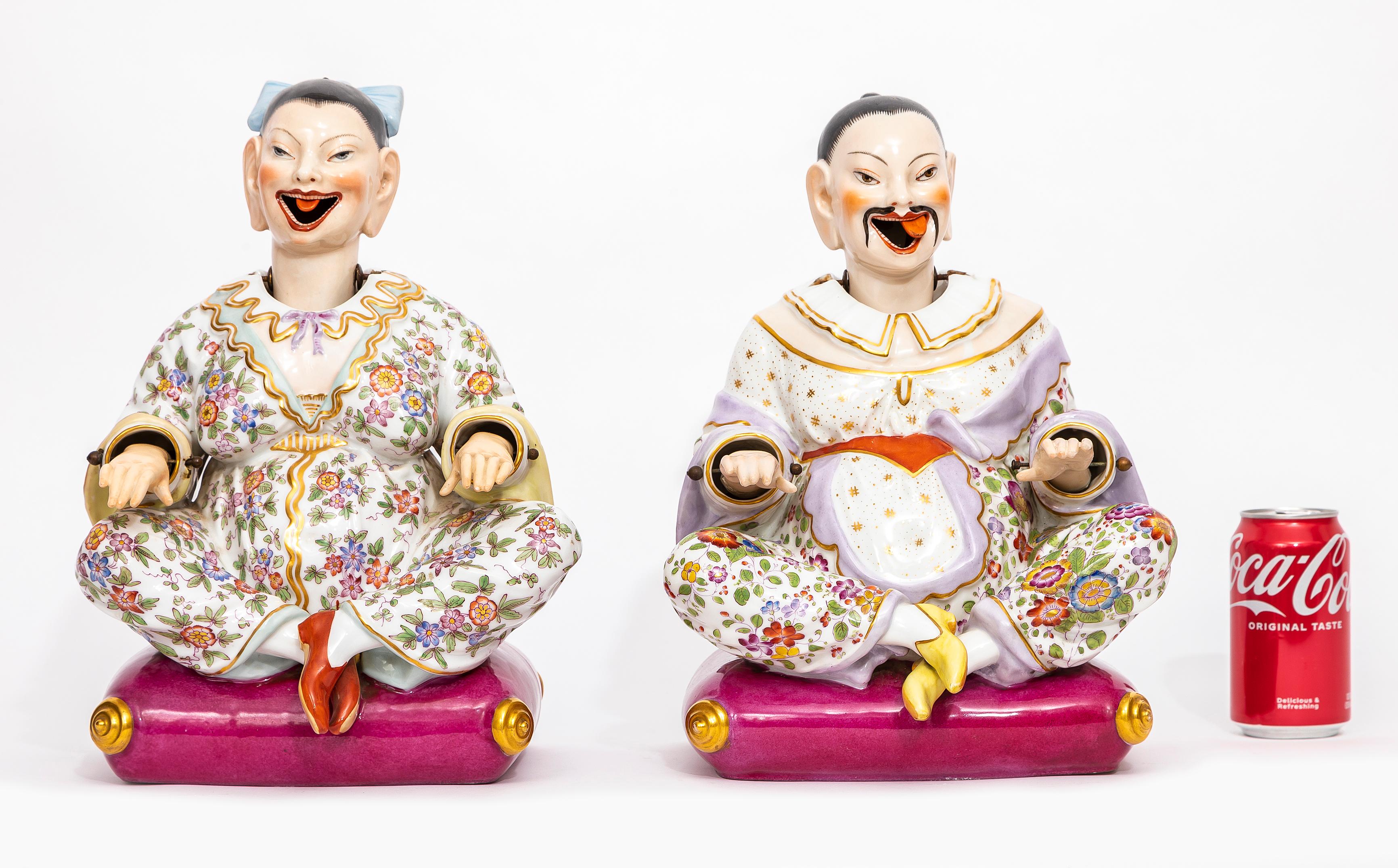Pareja de figuras de cabeza asintiendo de porcelana de Dresden del siglo XIX.  Cada figura está sentada con las piernas cruzadas sobre una vibrante base magenta con pies de pergamino dorados, diseñada con cabezas, manos y lenguas articuladas que