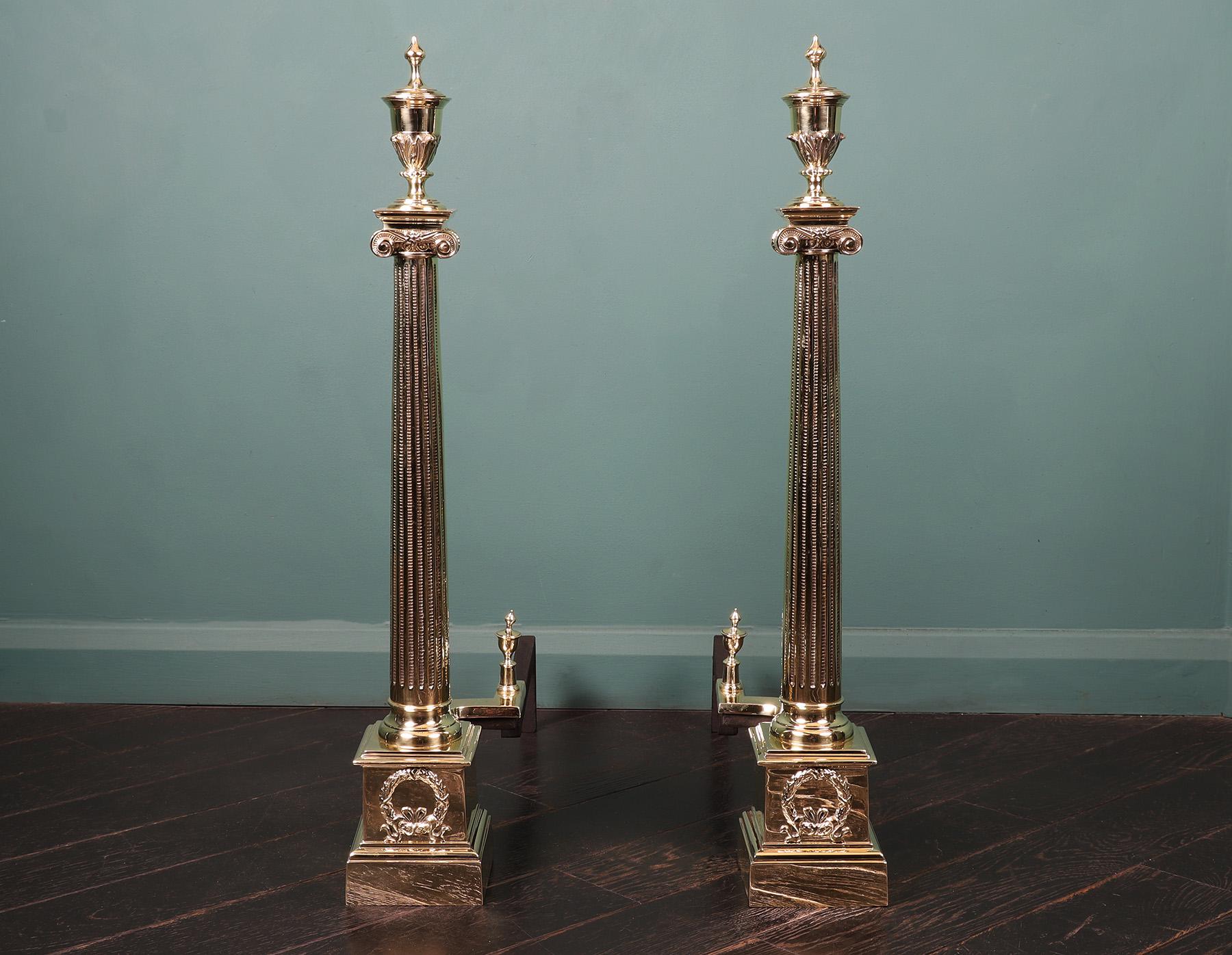 A pair of 19th Century Neo-classical brass andirons with fluted tapered ionic columns mounted on square plinths decorated with circular wreaths. The rear log-supports are in iron with overlaid section in brass, small brass urn finials uppermost.
