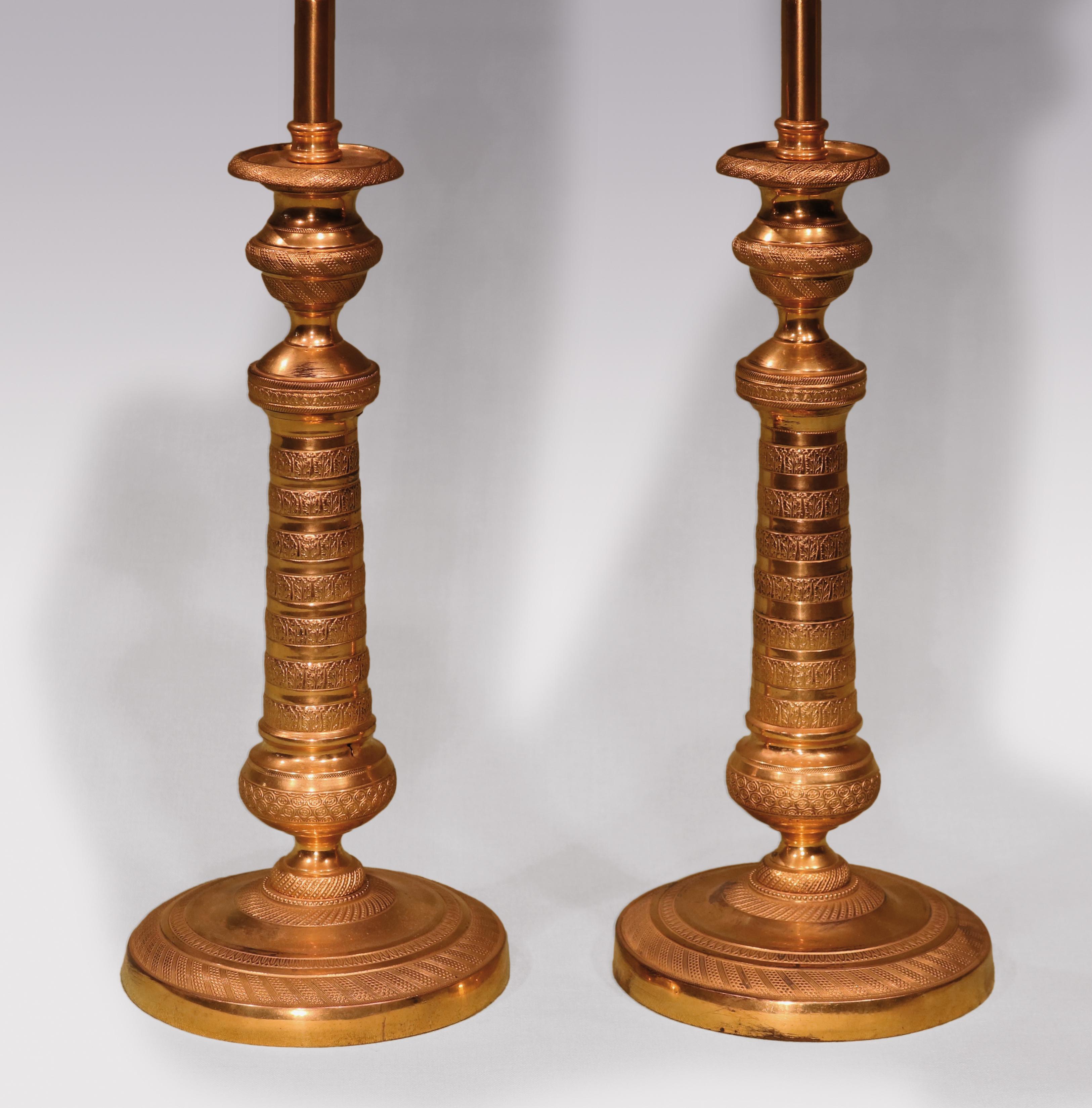 A pair of early 19th century ormolu candlesticks finely engine-turned throughout, having urn shaped nozzles above tapering stems ending on circular bases. (Now converted to lamps)

Measure: Height of candlesticks 27 cm (10.5 ins).