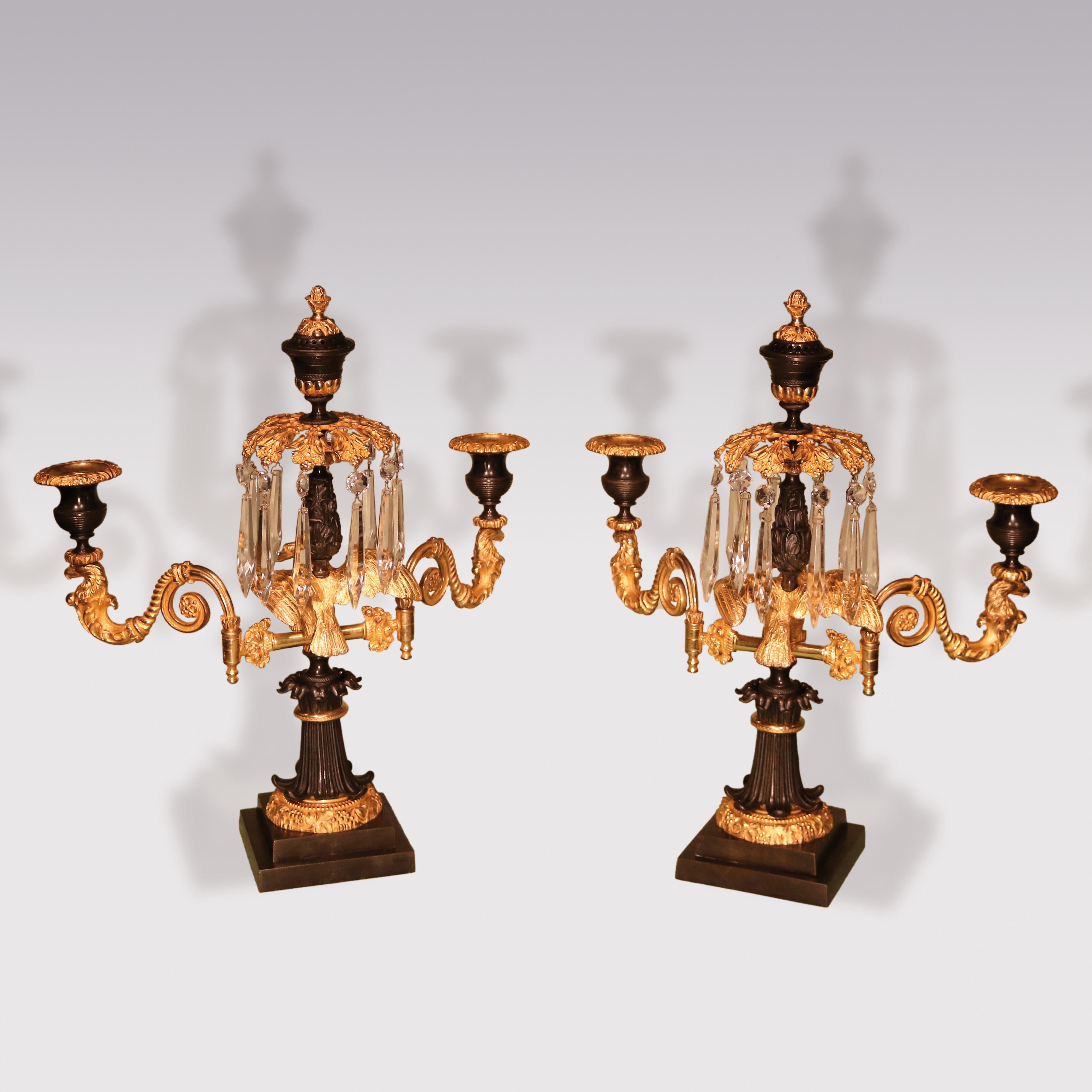 A pair of early 19th century Regency period bronze and ormolu 2-light lustre candlesticks having central urn finial above perched winged eagles, flanked by scrolled eagle-head candlearms, raised on leaf scroll columns with leaf and berry decoration