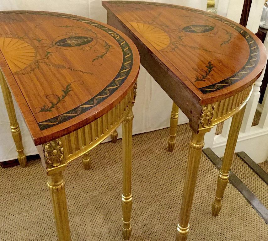 A pair of demilune satinwood and gilt console tables, circa 1920-1930. The tables are in a Sheraton style with painted foliage to the top. A twelve sectioned fan shell design is to the top and centre; below that is an oval shape with an urn and