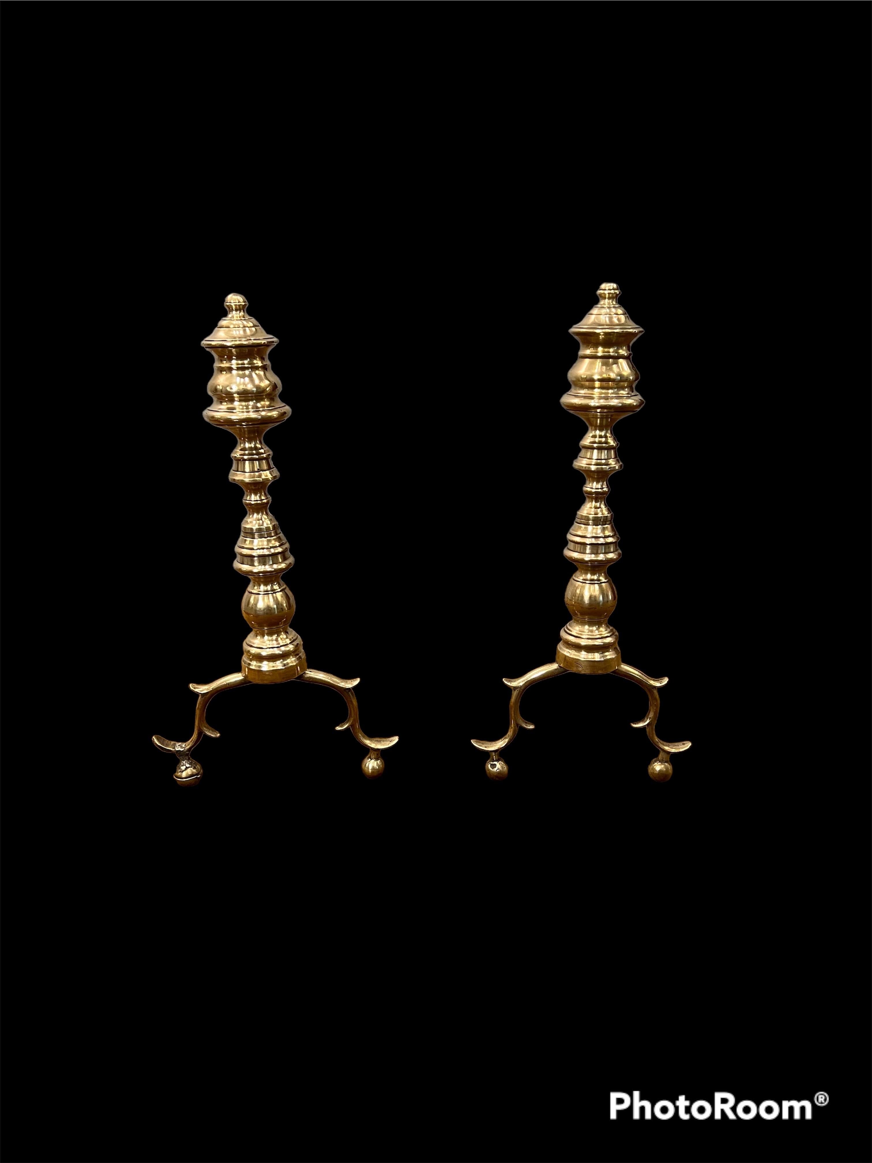 A Pair of American Federal Period finial form brass andirons, both showing turned finial capitals on sectional columns rising from double spurred and hipped supports, both retaining their original forged iron 'dogs',

The brass having a fine