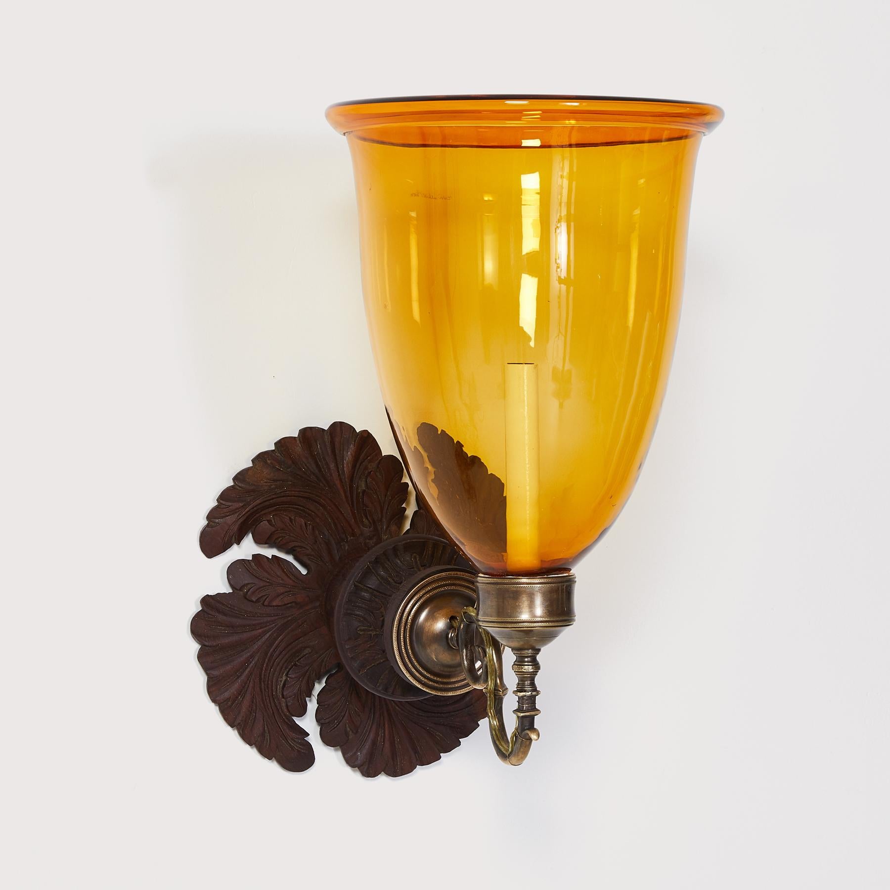 A pair of early 20th century hand-carved mahogany Anglo Indian styled backplate with patinated brass sconce fittings and rare hand blown amber glass hurricane shades. Electrified with one candelabra base socket per sconce.

Our custom hurricane