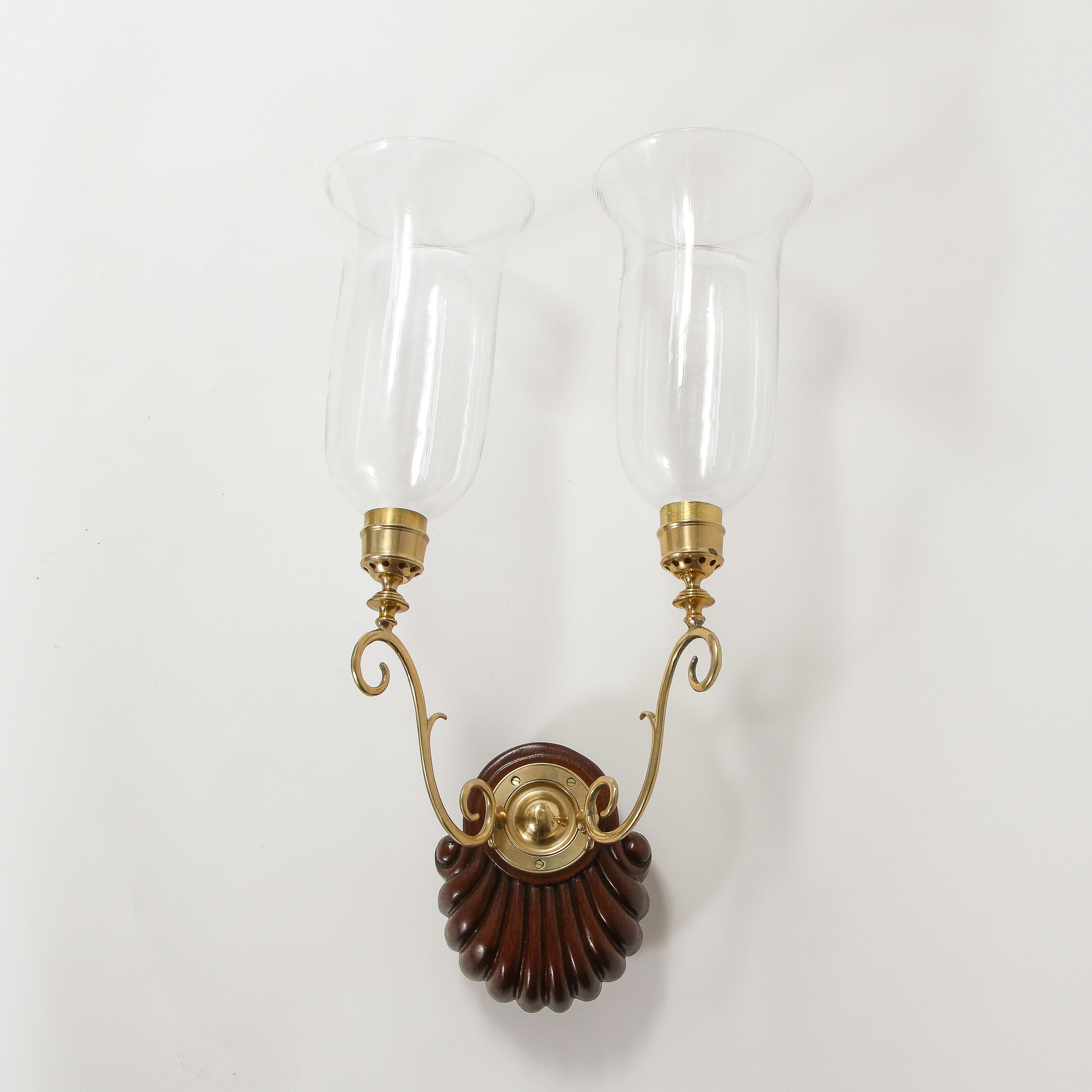 Each with carved hardwood shell-form backplate issuing two brass outward scrolling candlers fitted with bell-shaped clear glass hurricane shades.