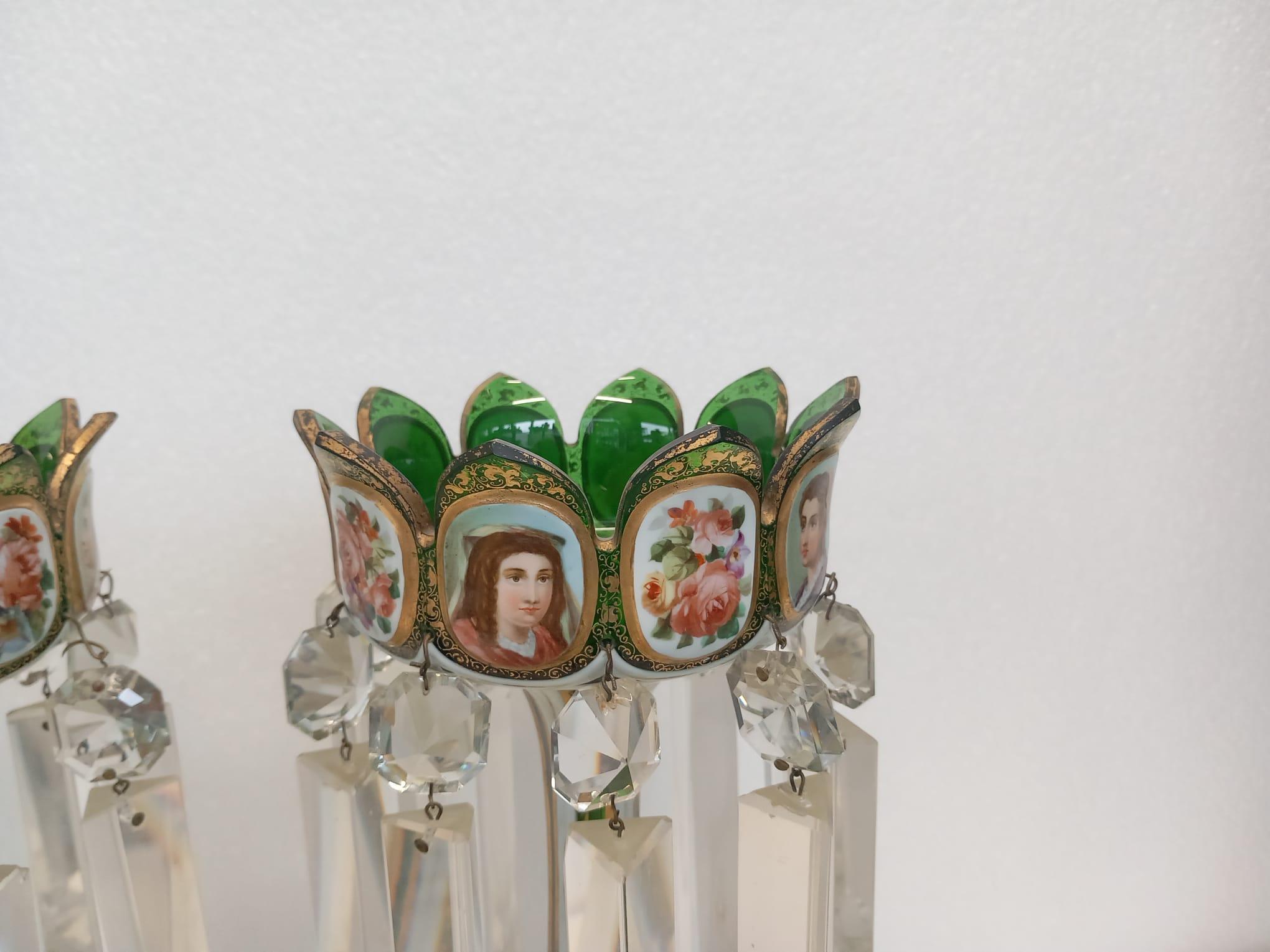 A pair of antique Bohemian green lustres with white and gilded cartouches, hand painted with court beauties and floral bouquets on a green glass background. The lustres are in clear crystal that hang over a gothic design white and gilt foot.
