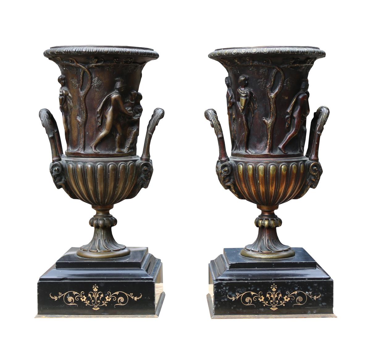 The Urns of campana form, each decorated with figures in classical attire with acanthus molded handles sat on black marble bases with scroll decoration to the front.