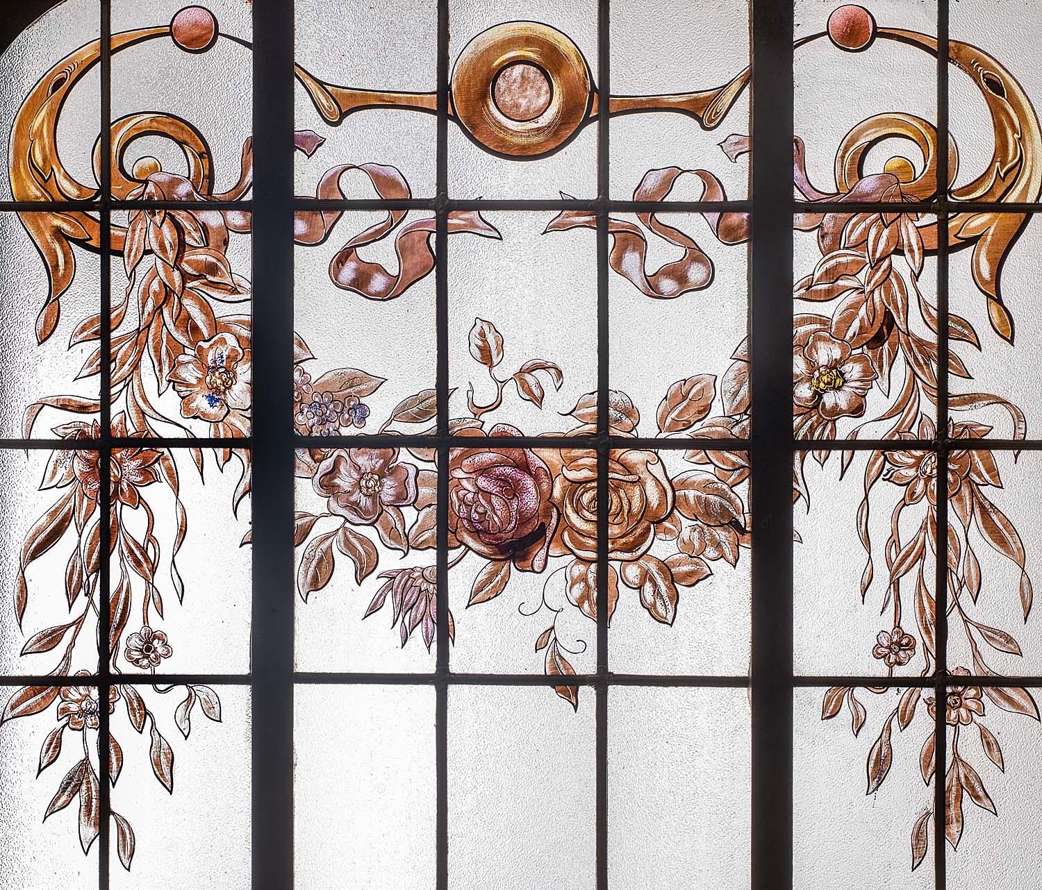 A pair of early 20th century, textured glass painted windows in rectangular leaded metal frames, decorated with naturalistic ribboned garlands of roses and foliage to the upper section above flamboyant sprays of flowers arranged within twin handled