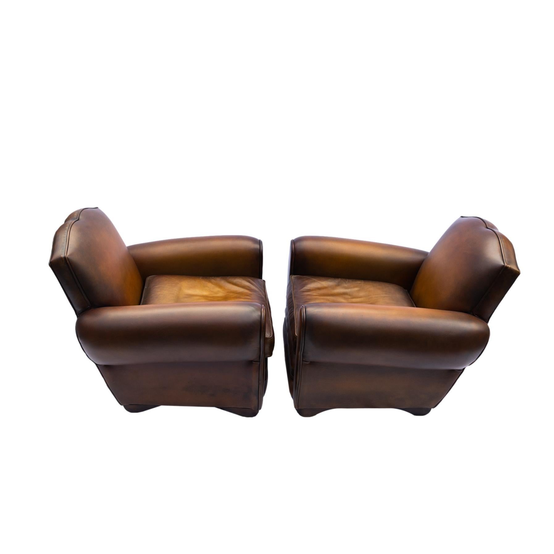 A Pair of Art Deco leather club chairs of Streamlined Modern Form, with rolled arms and arched sides, trimmed with leather welt cord, the mustache-shaped backs trimmed with nailheads, the decking detailed with thick leather piping, on oak block