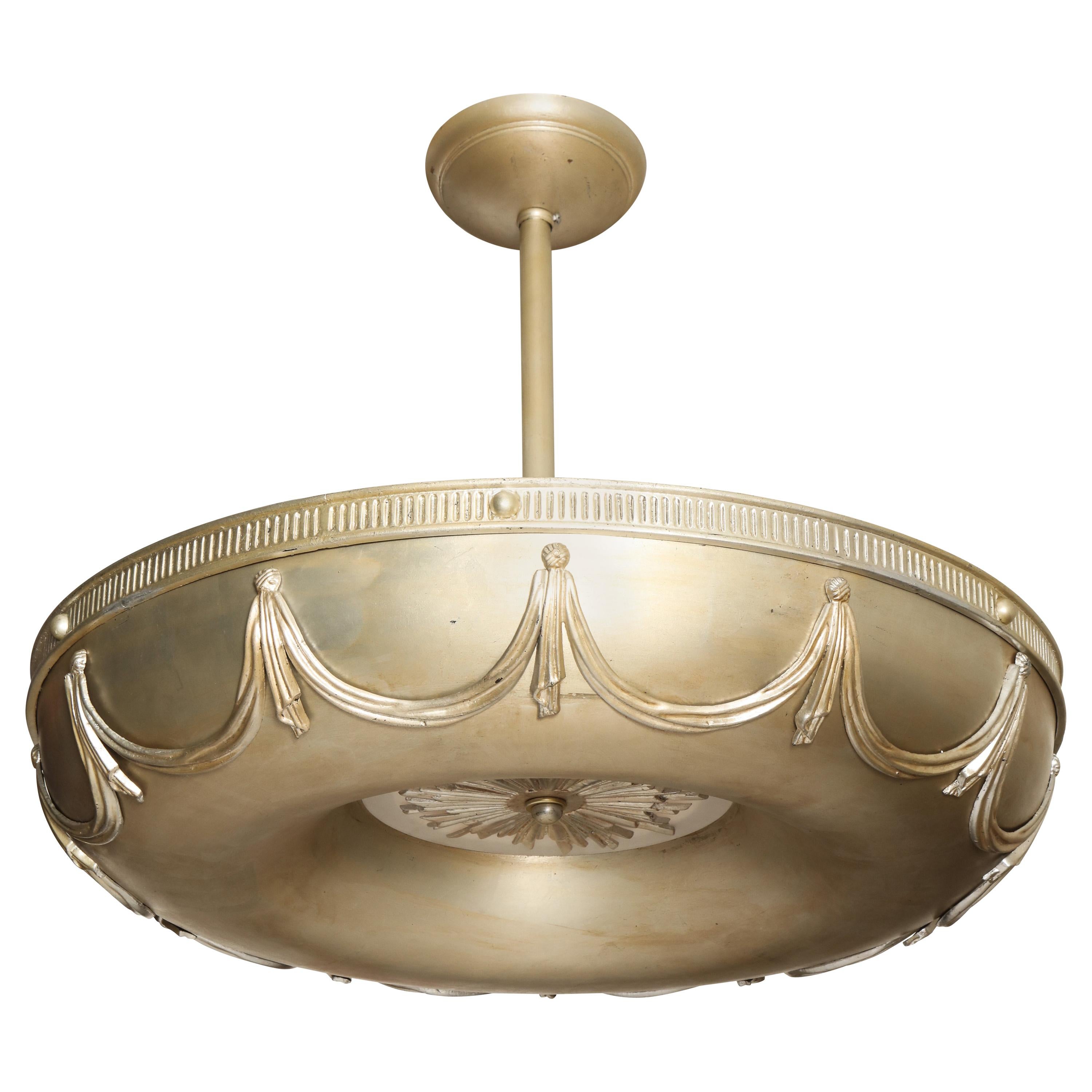 A pair of antiqued silver-leaf Art Deco, inspired chandeliers, with torus shaped frames and swag motifs. Each chandelier features a cast starburst shaped element on the underside with a piece of round frosted circular glass. Seven sockets under the