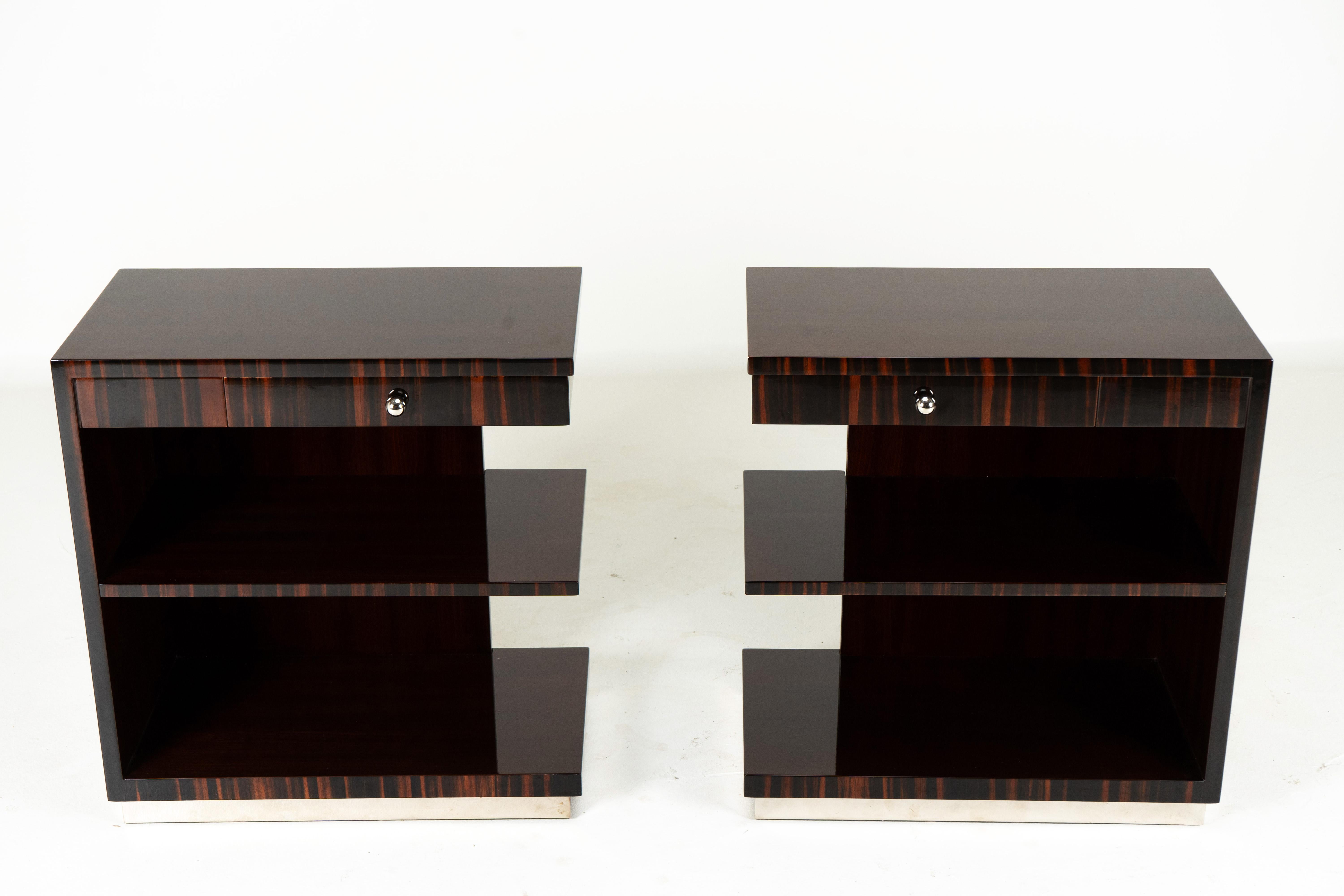 A Striking pair of Art Deco-inspired side tables in glossy walnut veneer. The tables feature 3 stacked and cantilevered shelves. The petite drawer fronts feature macassar veneer running vertically. At the bottom, there is a shelf for books or extra
