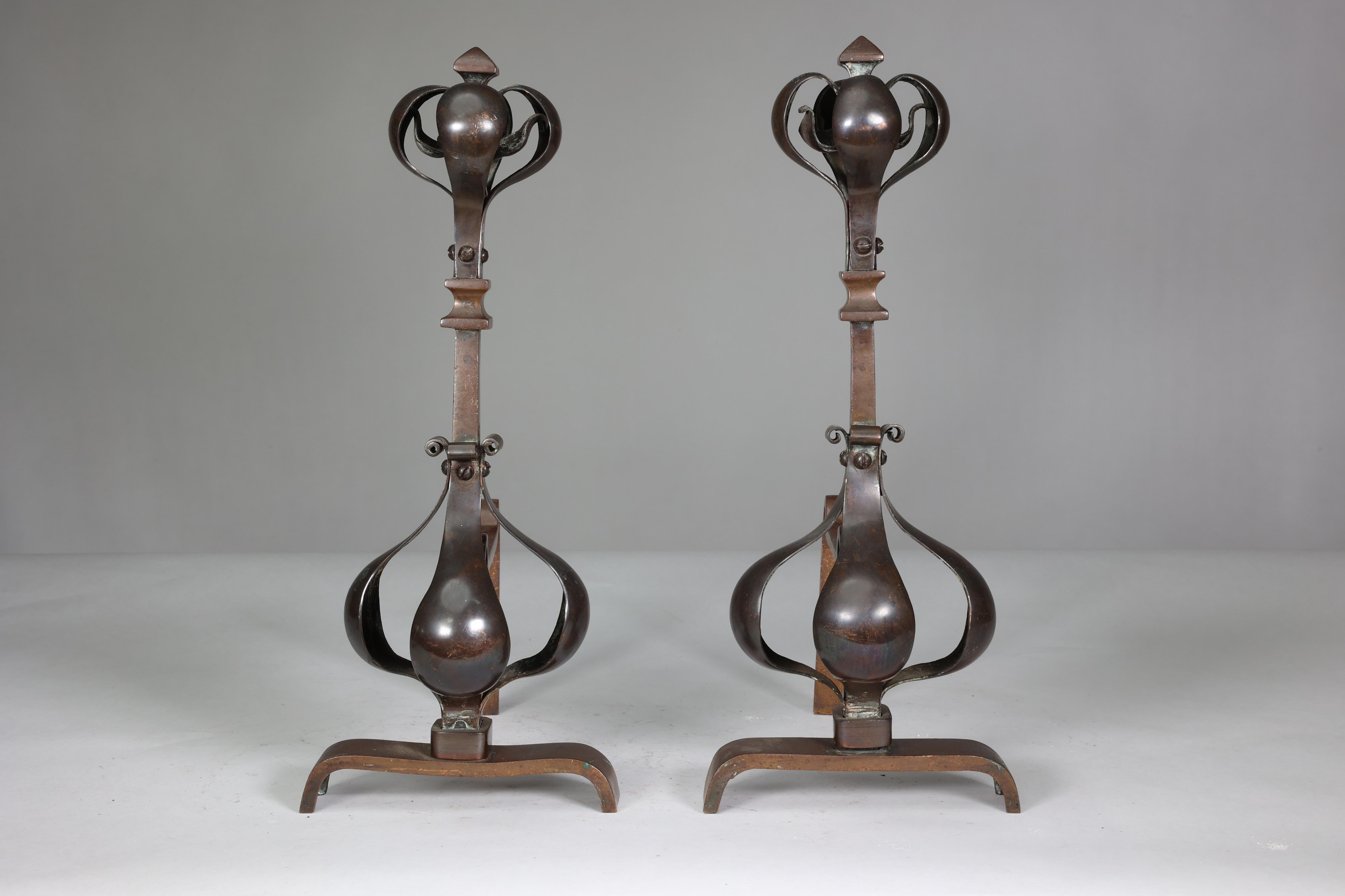 A pair of Arts and Crafts copper and bronze fire dogs with pyramid finials and decorative with copper flower heads to the tops, and copper flower decoration to the bases applied to a bronze frame.