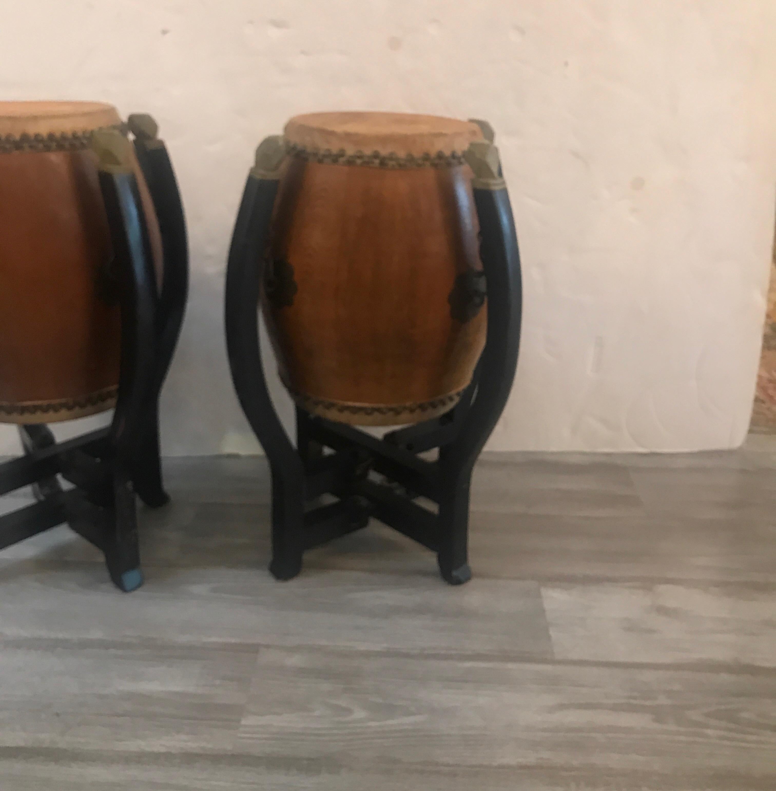 A pair of barrel shaped drums with a drum surface on the tops and bottoms with nailhead trim around the animal skins. The bases in original black paint that fold flat. Some old restoration on one leg.
