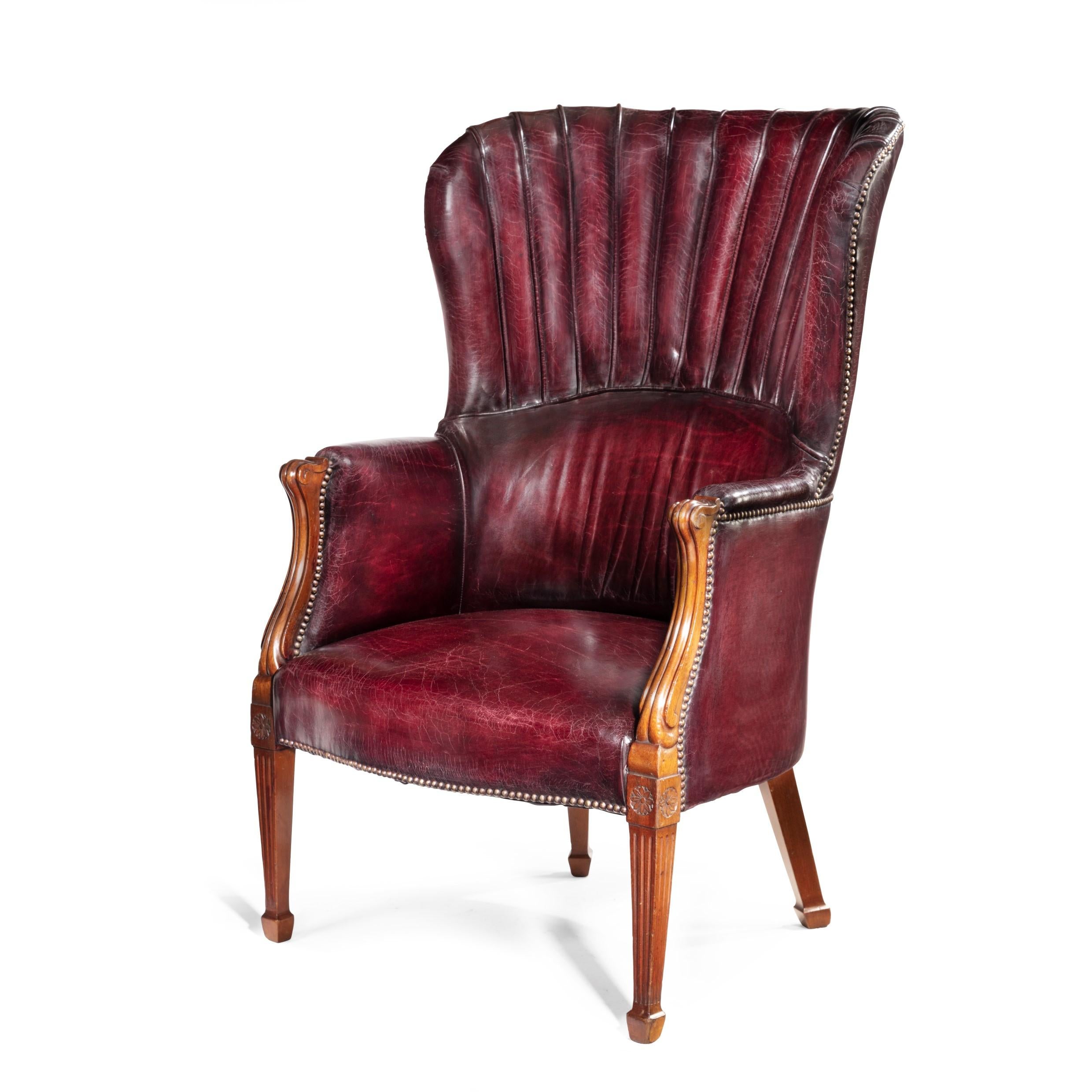 A pair of barrel-backed mahogany wing armchairs, each with carved serpentine arms and fluted square section tapering front legs with spade feet, the rear legs out-swept, reupholstered in distressed red leather. English, circa 1900.