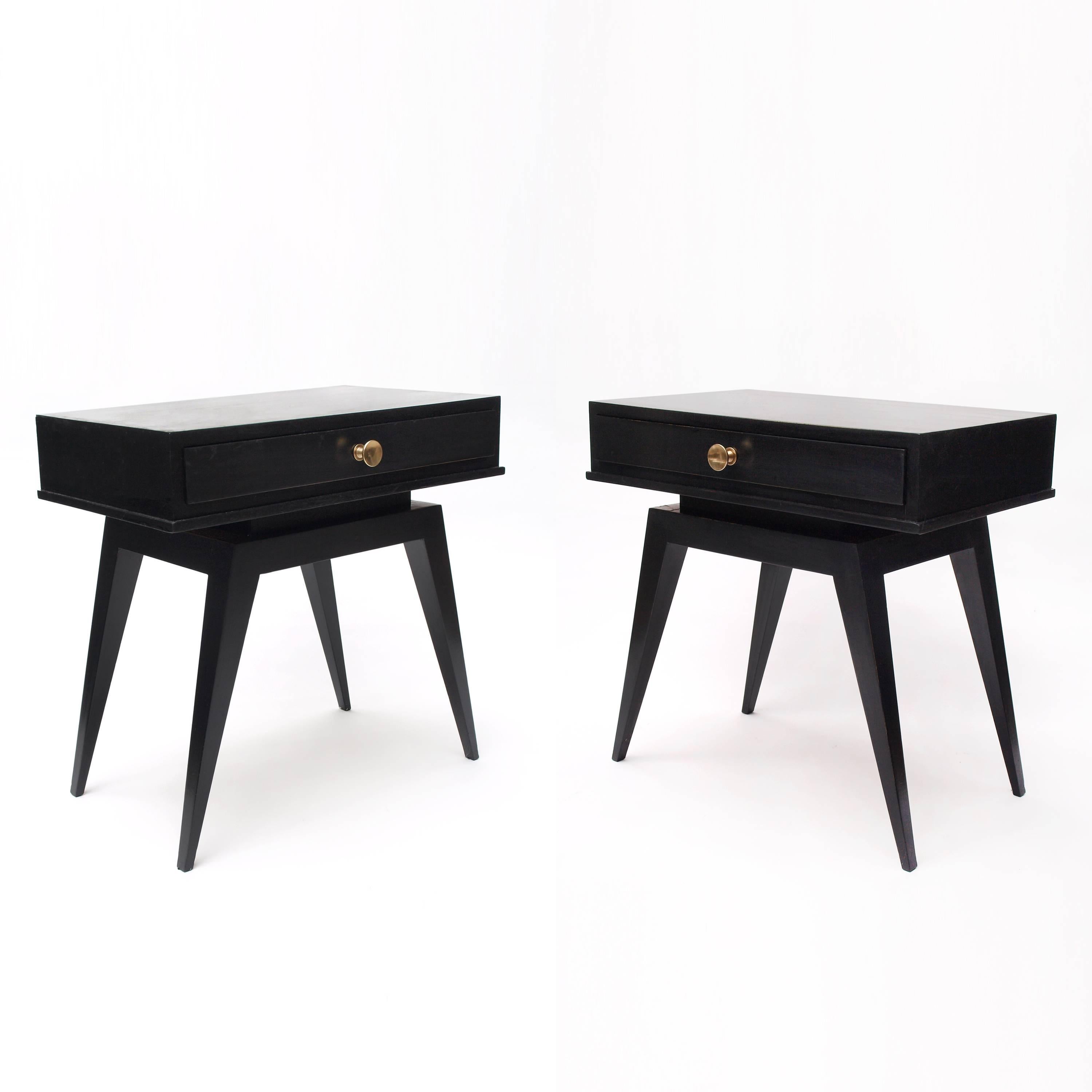 A pair of black 1940s French nightstands. Each stand has a single shallow drawer adorned with a brass handle featuring a subtle conical concentric motif. The drawer section is supported on a staggered plinth to appear floating above the