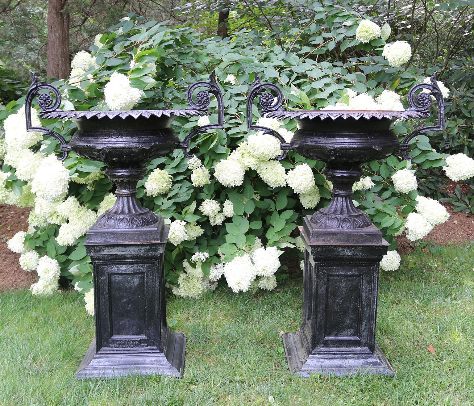 A pair of cast-iron “Poplar” urns on pedestals by J.W. Fiske Iron Works, the urns with arabesque handles with rosette flourishes and pointed scalloped rims, the bodies of the urns with acanthus leaf motif, and the fluted socles with modified egg and