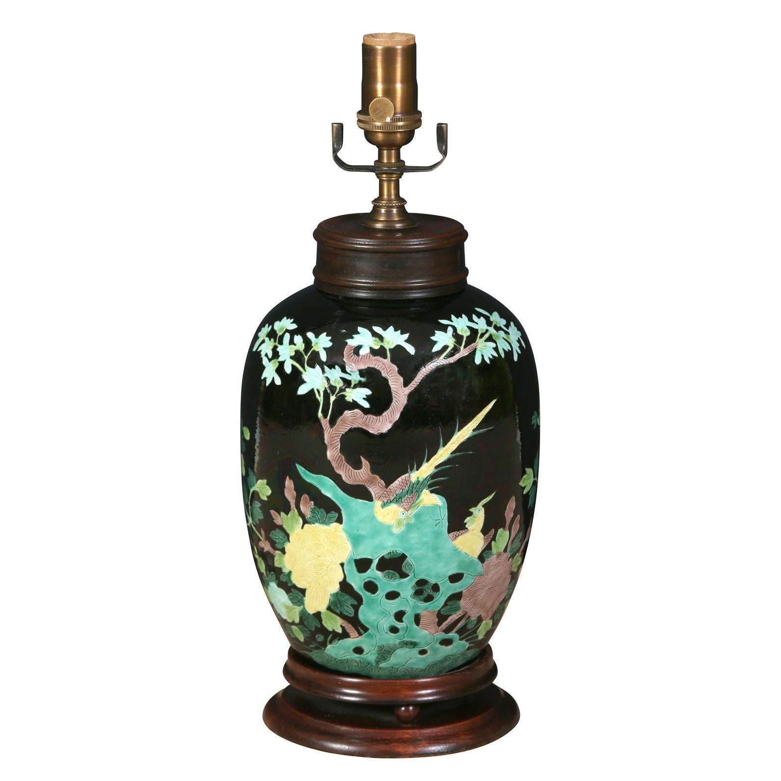 A pair of eye-catching Chinese export porcelain lamps with a black ground, decorated with branches, flowers and butterflies in beautiful tones of green, yellow and soft brown. The lamps are urn shaped and are mounted on a stained wood base.