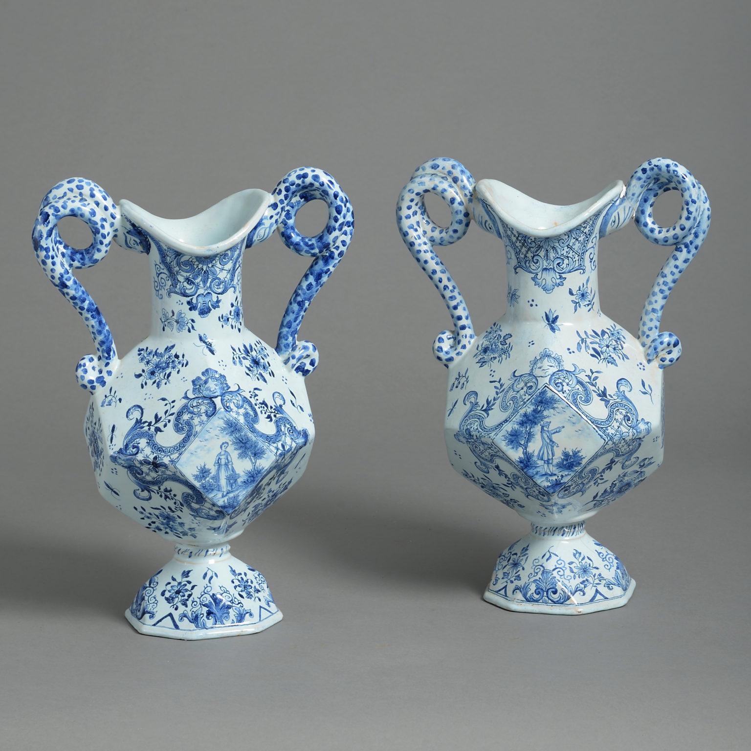 A pair of late 19th century blue and white glazed faience pottery campagna vases, having scrolling handles, the bodies of faceted form and decorated with figurative scenes and armorials.