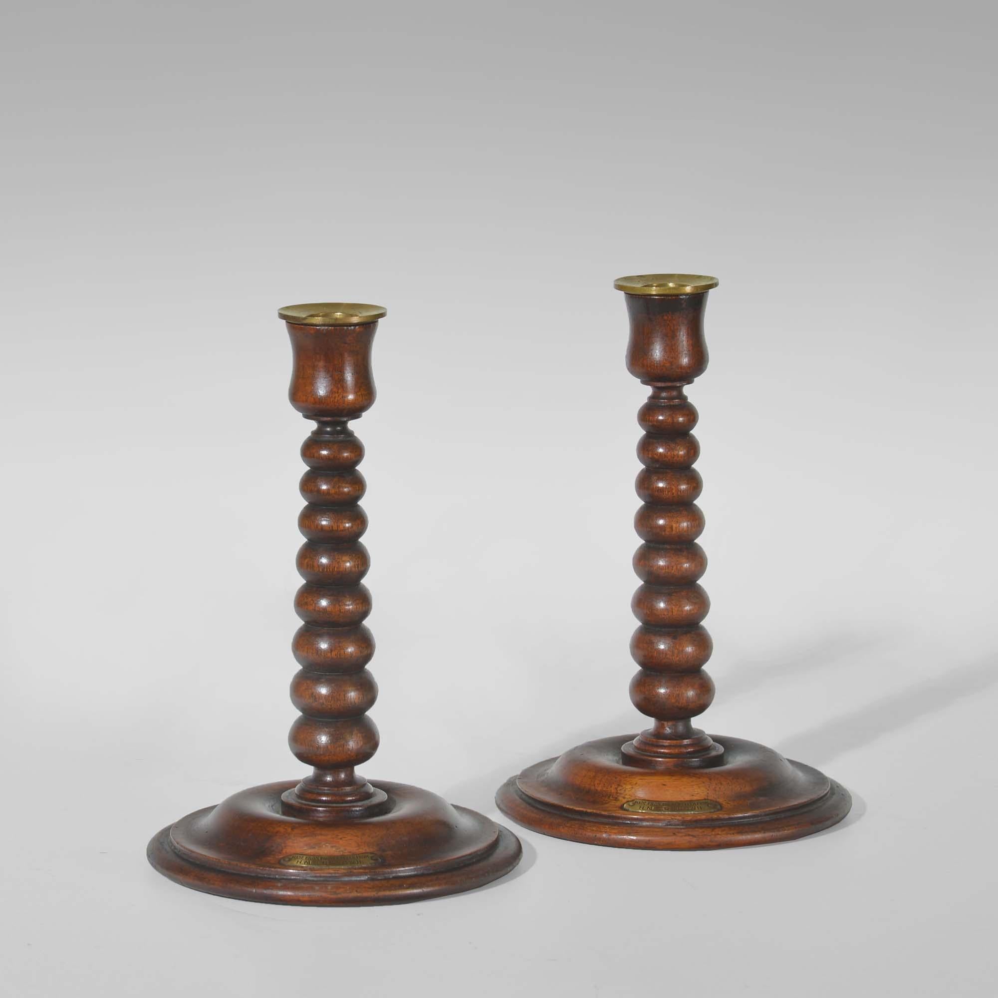 A pair of bobbin turned teak candlesticks - Royal yacht HMS Renown
 
A pair of teak candlesticks. The brass drip pans to concave turned candle sockets over tapered bobbin turned columns and moulded domed circular bases. Bear brass labels ‘Made from