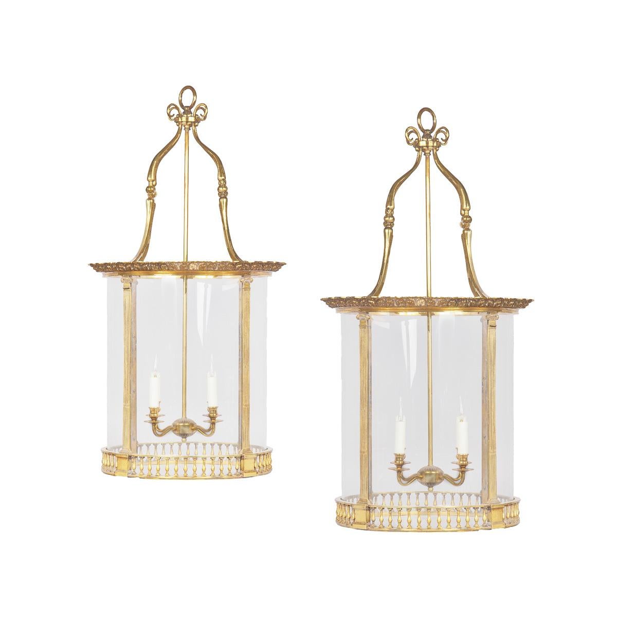 A pair of brass drum lanterns in the style of Lutyens, each of cylindrical form with four candle-arms suspended inside on a single tubular support, the four curved glass plates held within a frame with an everted shaped rim embossed with floral