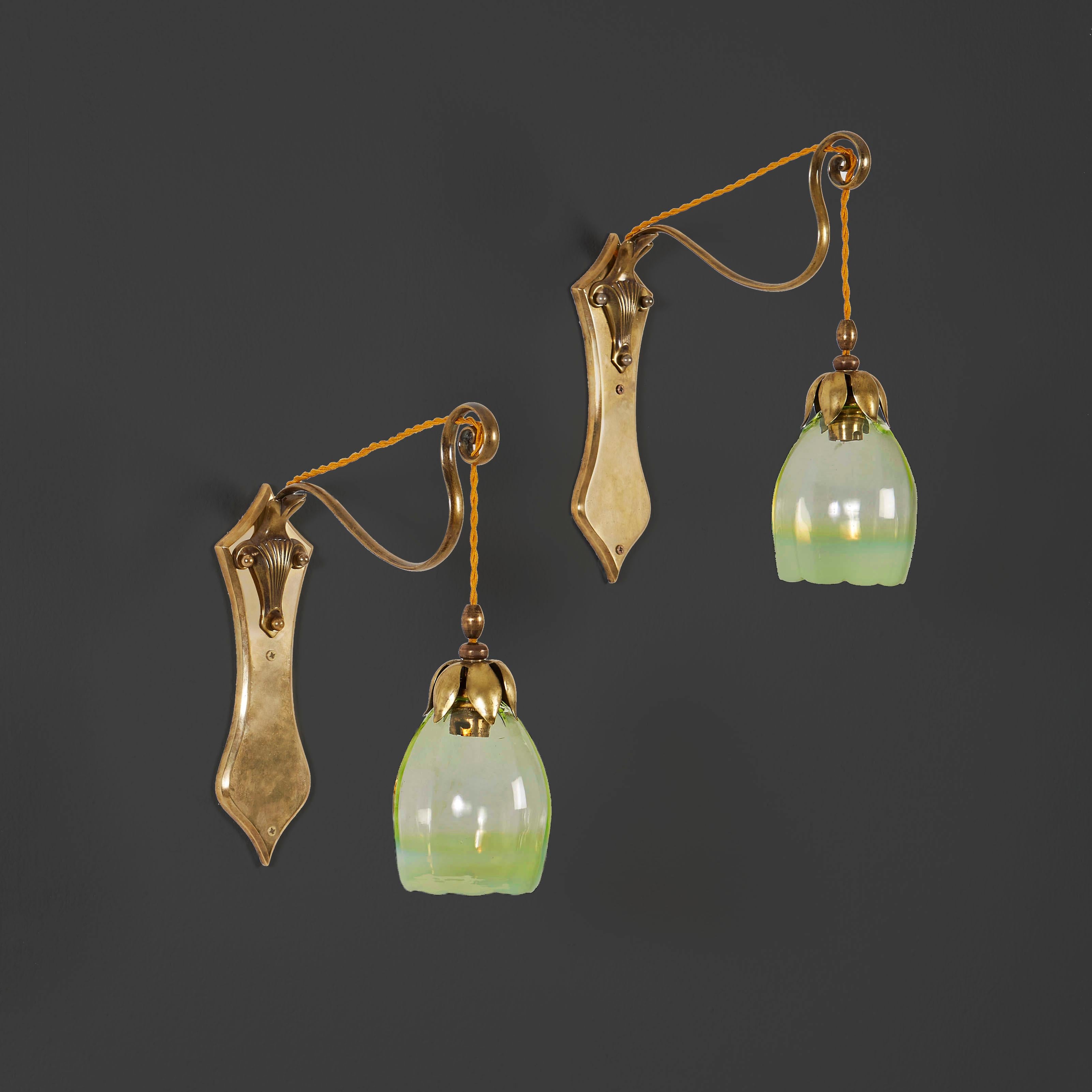 A pair of wall lights by W. A. S. Benson, with scrolling arm supporting the original opaline glass shade in the shape of a flower, decorated with brass sepals, all mounted on brass plates decorated with abstract foliate motif.

Currently wired for