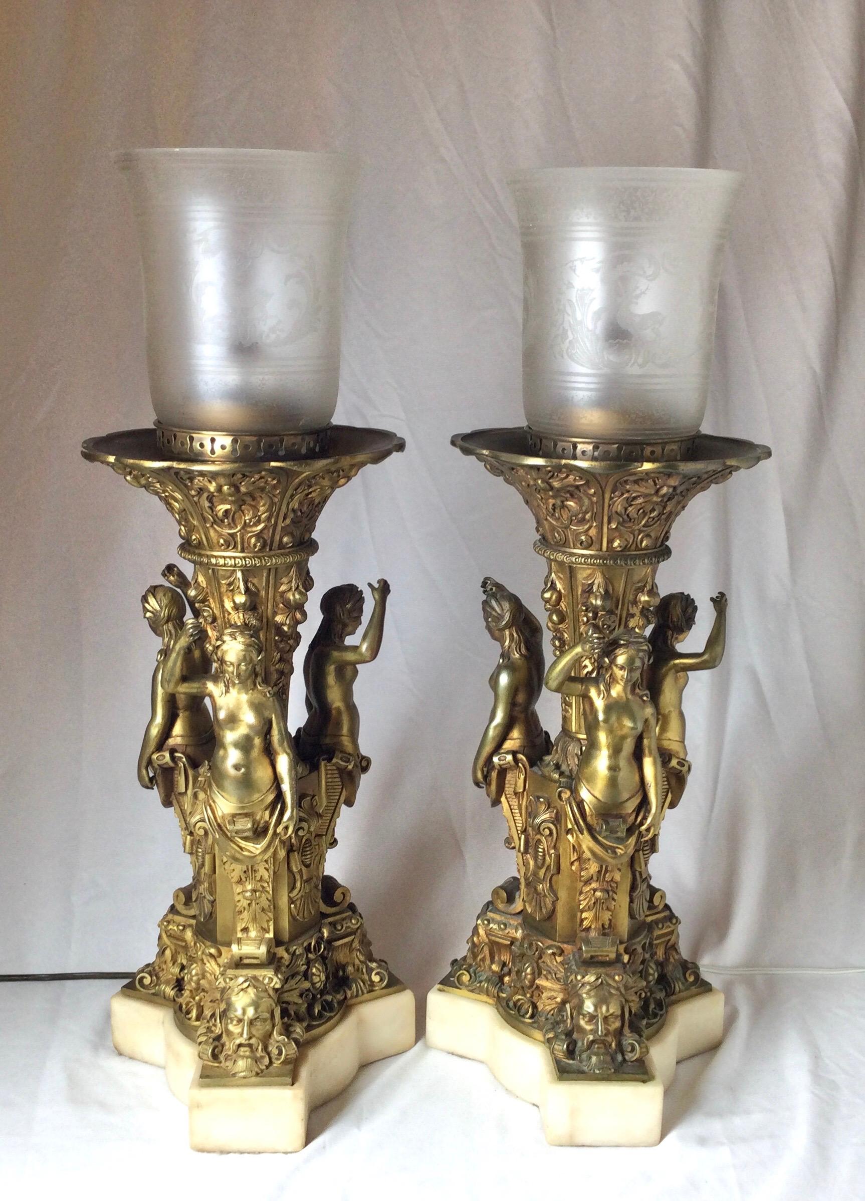 A pair of gilt bronze figural neoclassical lamps with marble bases. The bronzes with gilt surface showing wear but original. The off white marble bases fitted to the bronze figures. The etched glass shades soften the bulb. These can be changed to a