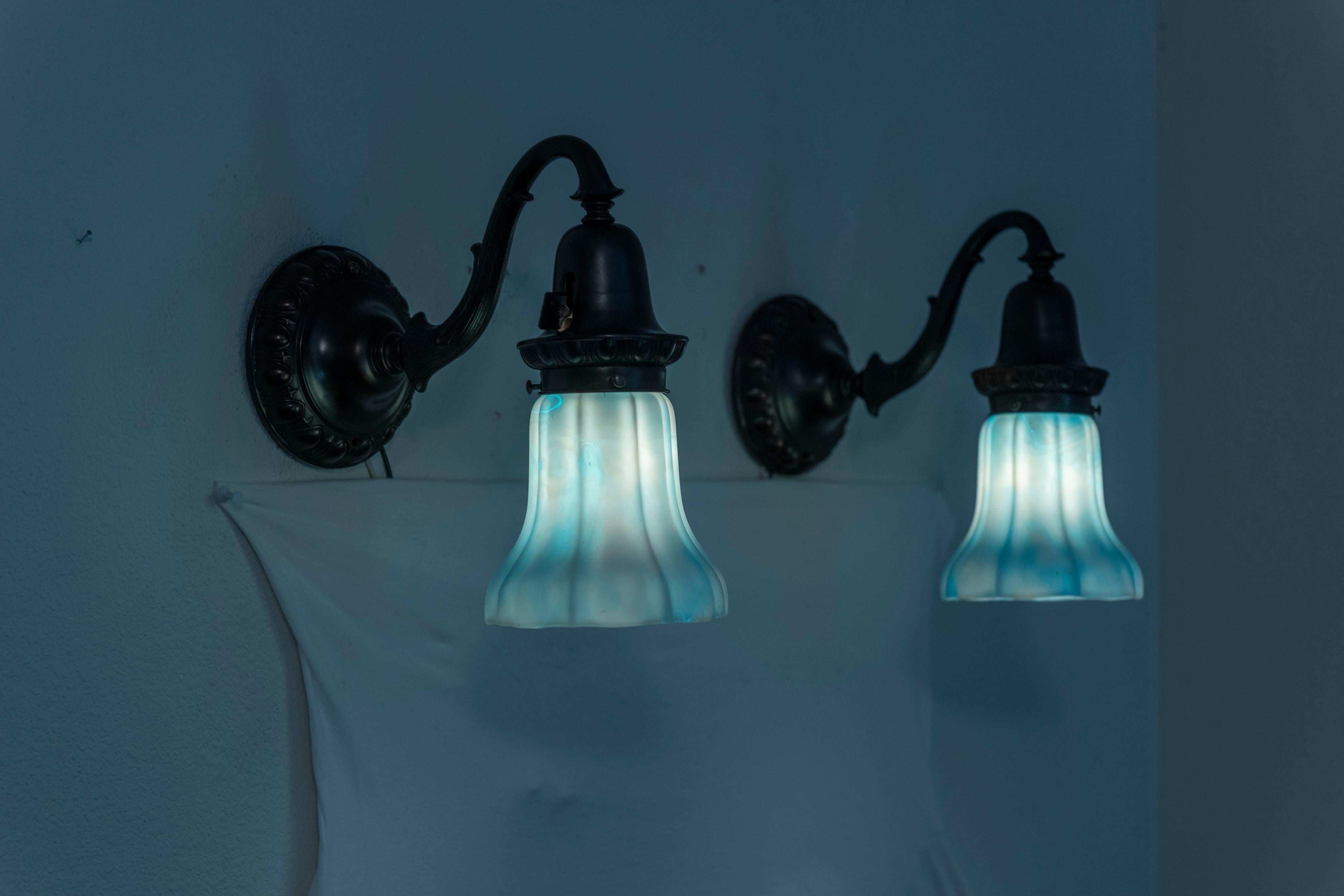 These warmly patinated bronze sconces are enriched with a pair of rare and wonderful Kokomo Co. glass shades. We have sold many items with glass by Kokomo, but this color of blue and white mixed together is an example we have never seen in our 40+