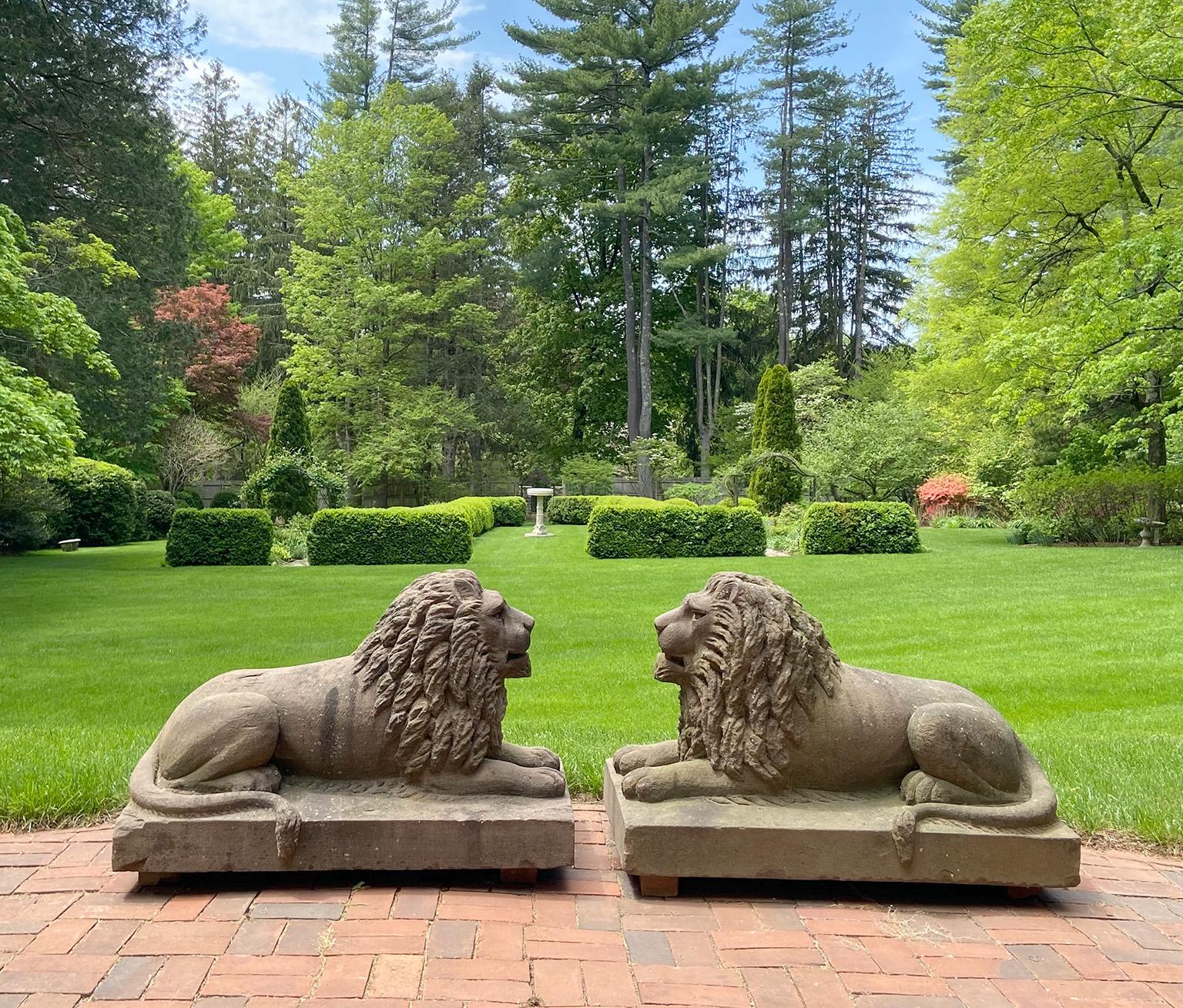 A rare pair of carved brownstone American Folk Art lions, in recumbent pose with full manes and tails curled to opposing sides, their expressive faces with mouths slightly open, accentuated by stippled whisker marks and soulful lidded