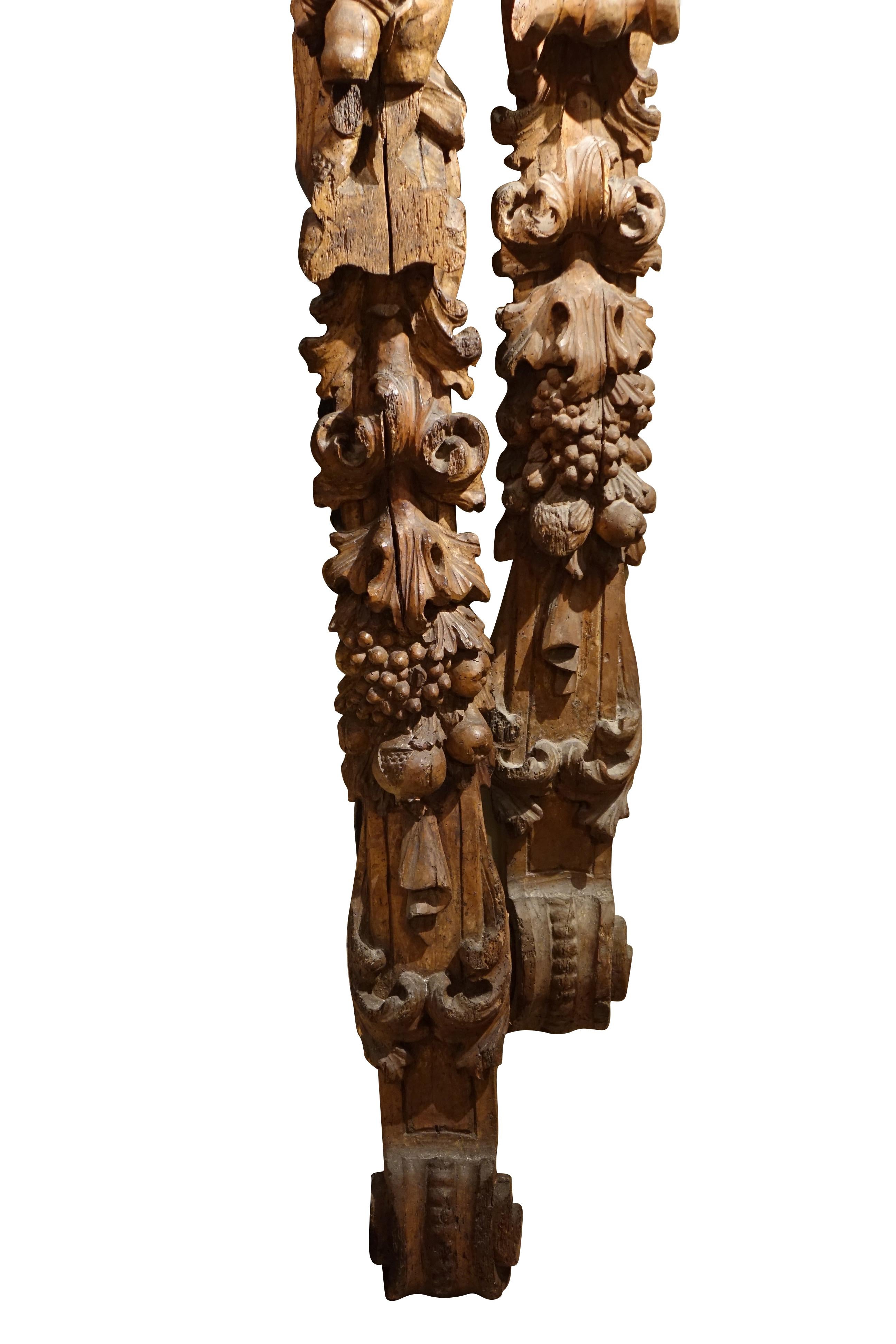 Pair of large carved wooden elements (1.90m), representing two puttis leaning against a cornice and supporting with one arm a scroll ending the whole (and forming the base of the composition).
The lower part is carved with curly leaves, bunches of