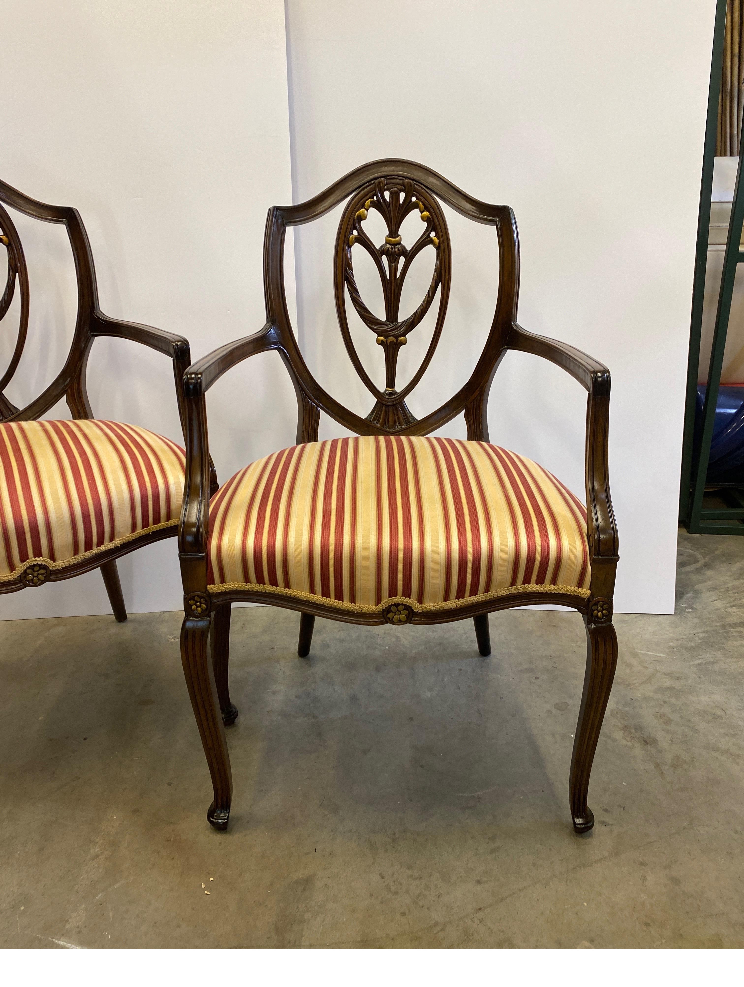 A graceful pair of hand carved mahogany Hepplewhite shield back armchairs. The backs with pierced shields with swags and gilt accents in the Prince of Whales motif with striped ribbon pattern satin seats in red, gold and cream. The front legs gently