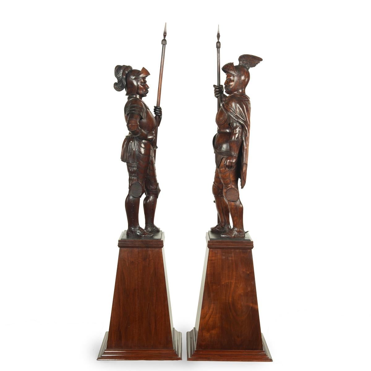 A pair of carved walnut mediaeval knights, each wearing full armour with a visored helmet metal, breastplate and holding a lance, standing on a later pyramidal base, one with curled feathers in his helmet and the other with twin ribbons, both with