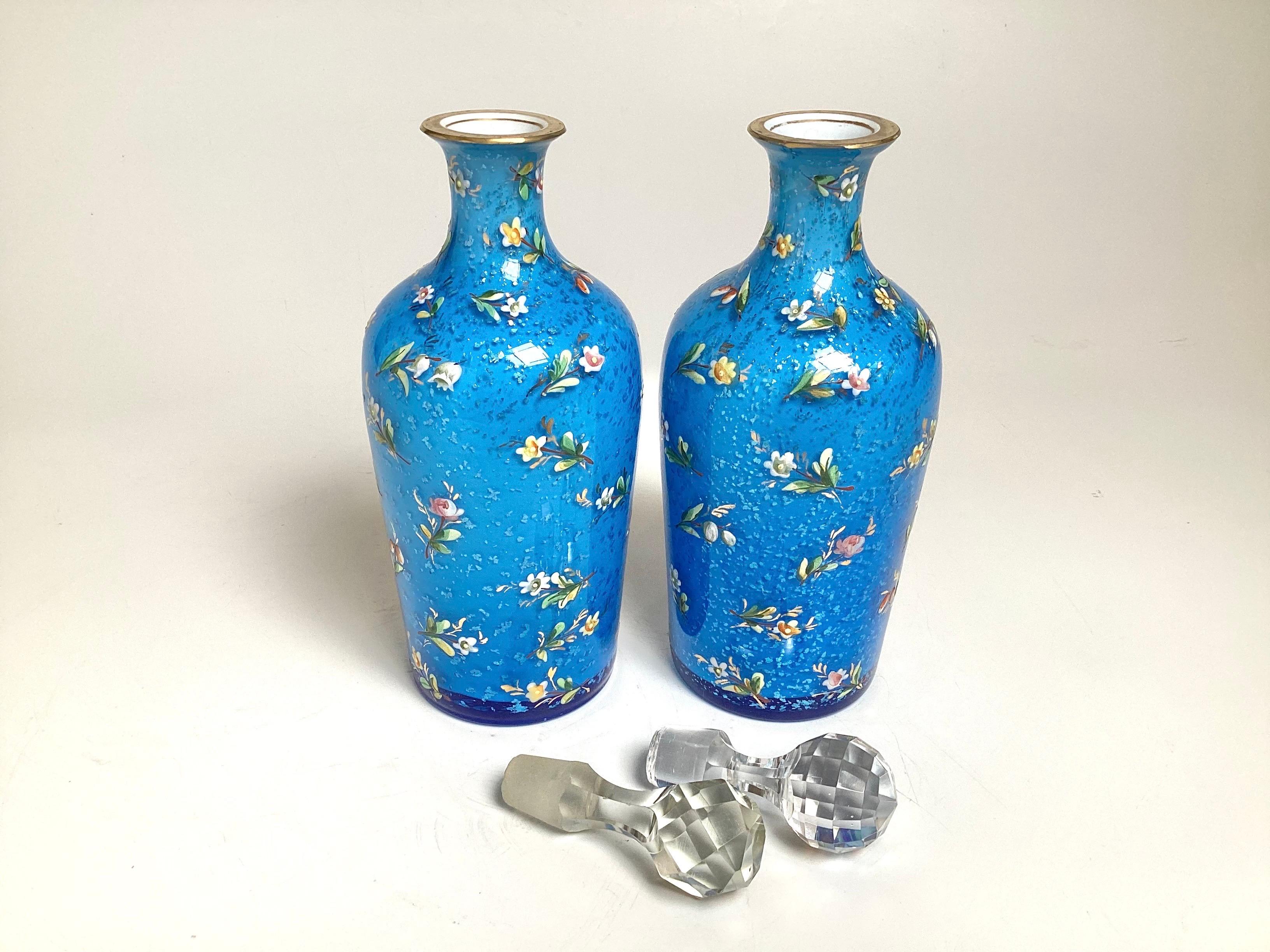 Una bella coppia di bottiglie di profumo in vetro blu ceruleo con decorazioni in smalto dipinte a mano. Le bottiglie presentano un motivo floreale all over con dettagli dorati nella parte superiore, i tappi sfaccettati nella parte superiore.  Un