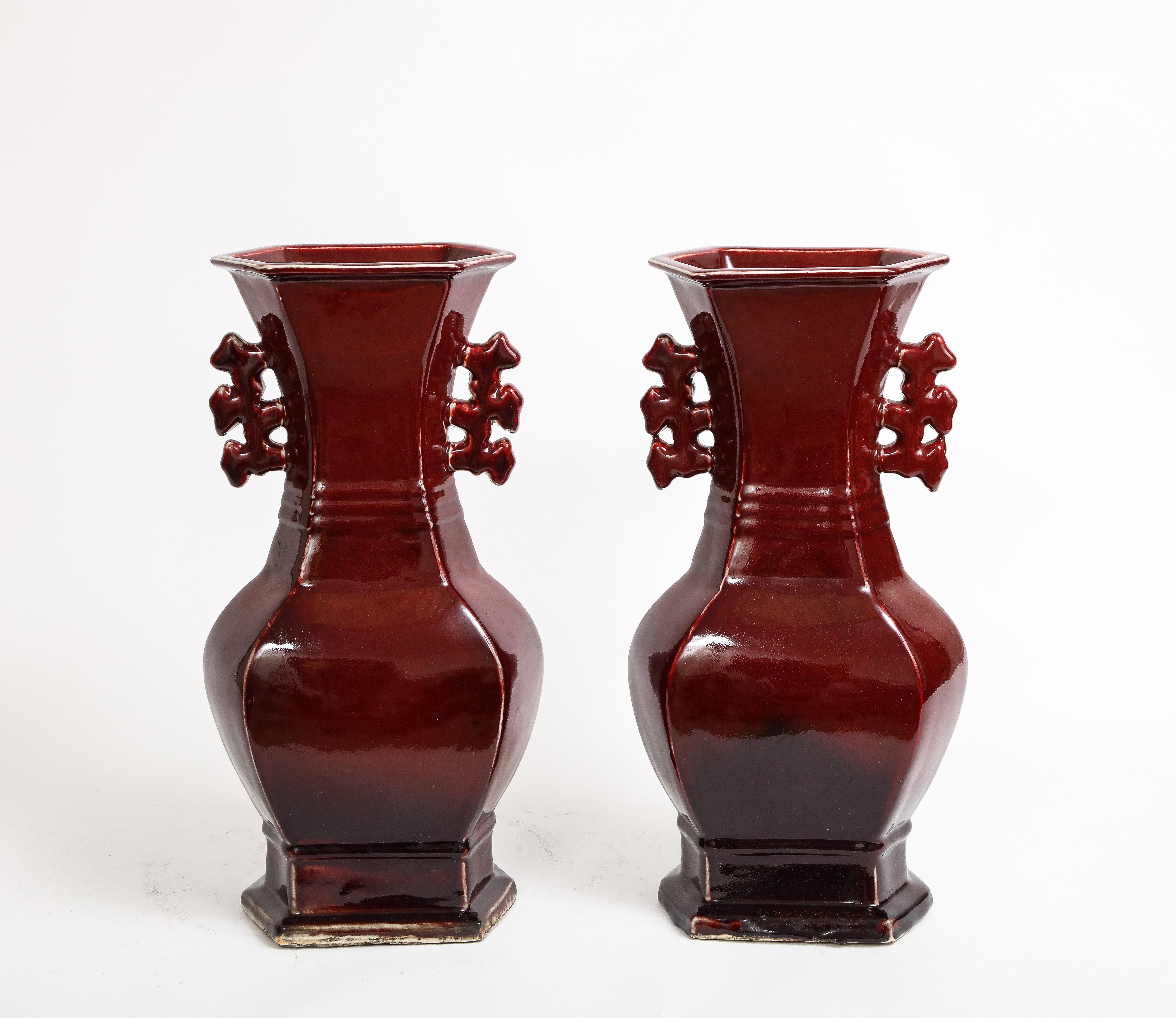 An Exquisite Pair of Chinese Monochrome Ox Blood Porcelain Double Handled Vases. The deep, rich hue of ox blood gradually transitions to a more translucent tone towards the upper portion, creating an alluring visual effect. These vases boast an