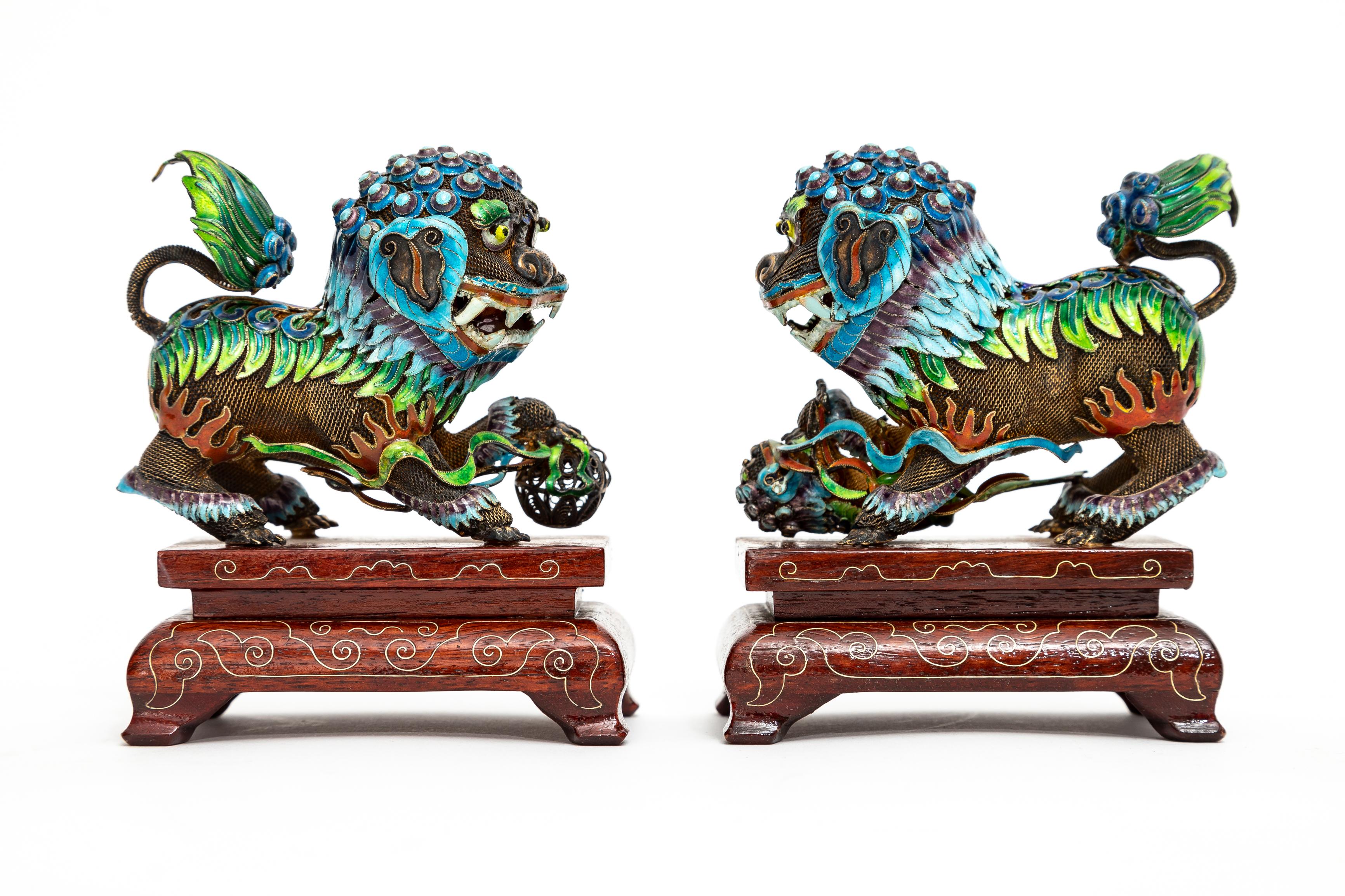 A Pair of Chinese Silver Filigree Enameled Foo Dogs on Carved Wood Stands.  Each foo dog is made up of very fine silver strands which have been weaved together creating an elaborate display of these foo dog objects.  One holds a ball underneath its