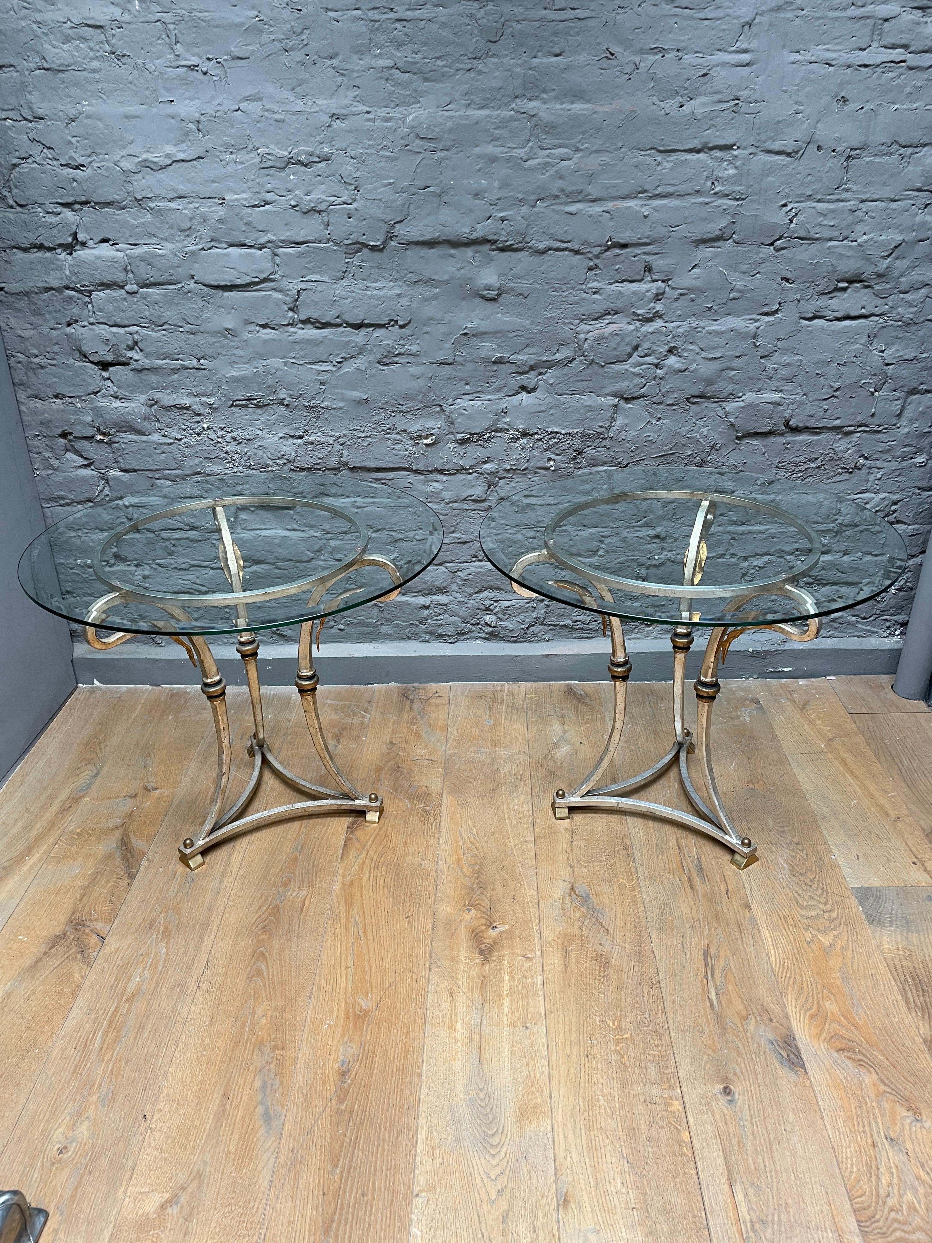 A circular pair of side tables with thick round bevelled glass tops, stood on brass block feet which support scrolled supports fishing in a gold gilt leaf. The frames in a nickel finish with gold leaf accents.