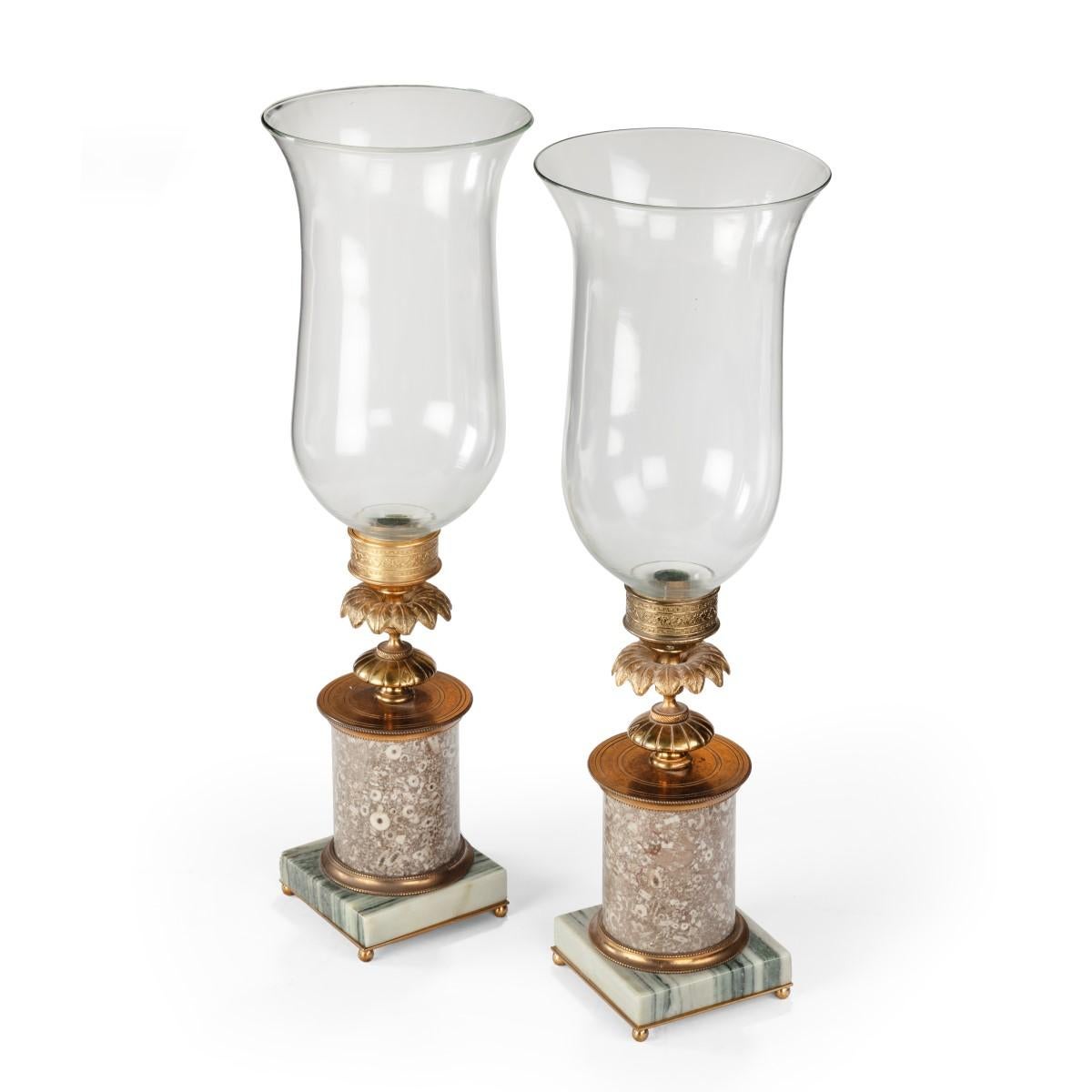 A pair of decorative storm lamps, each with a candle holder enclosed in a clear glass shade set on a turned gilt-brass support with a collar of petals and a cylindrical marble column, the rectangular base of striped marble. English, early 19th
