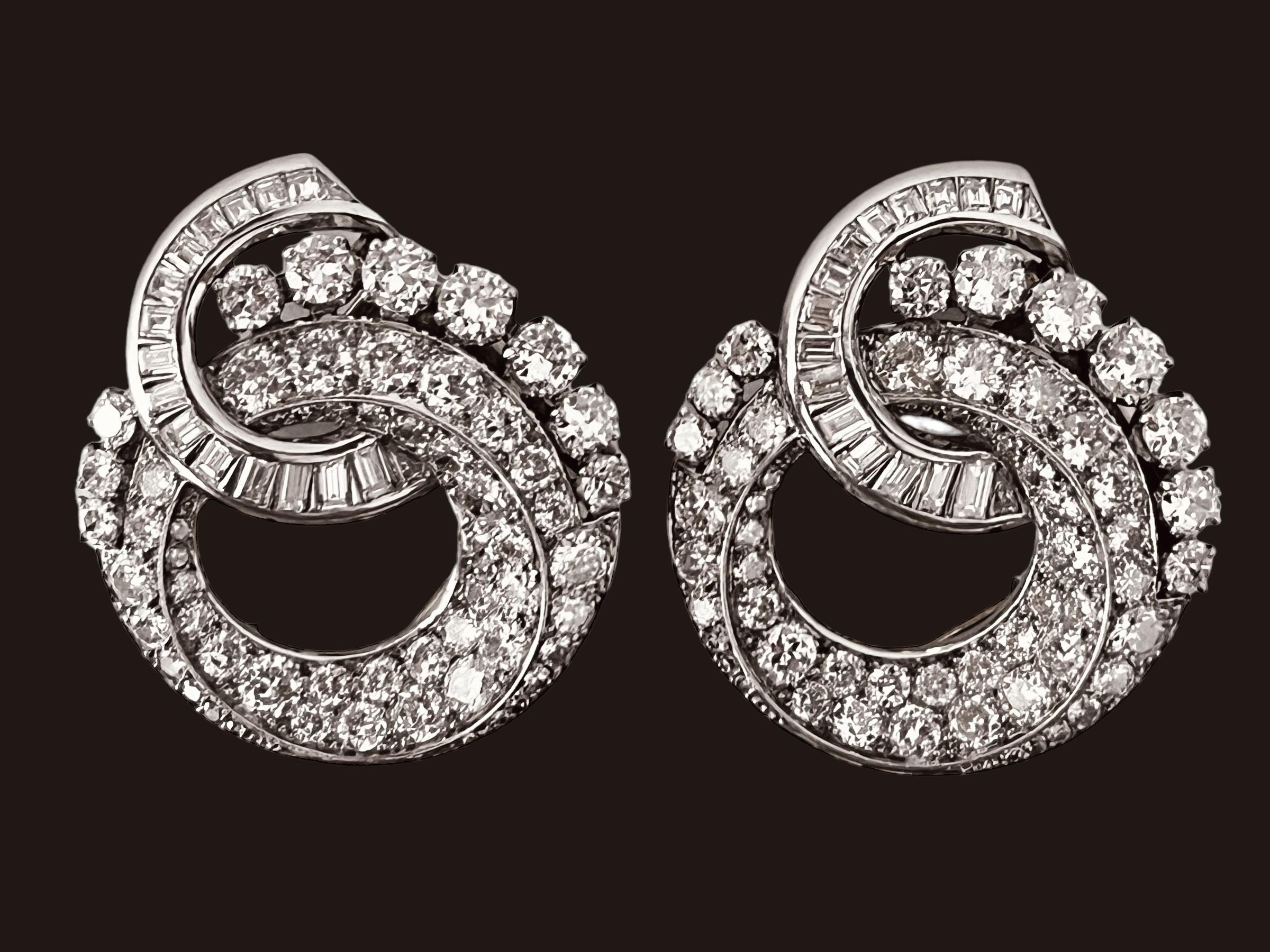 A pair of diamond clip brooches, each of swirling circular design. Claw and pave'-set with brilliant cut diamonds, accented by a channel-set baguette-cut diamond ribbon. Approximately 10cts of diamonds. Mounted in platinum. Circa 1930. One clip