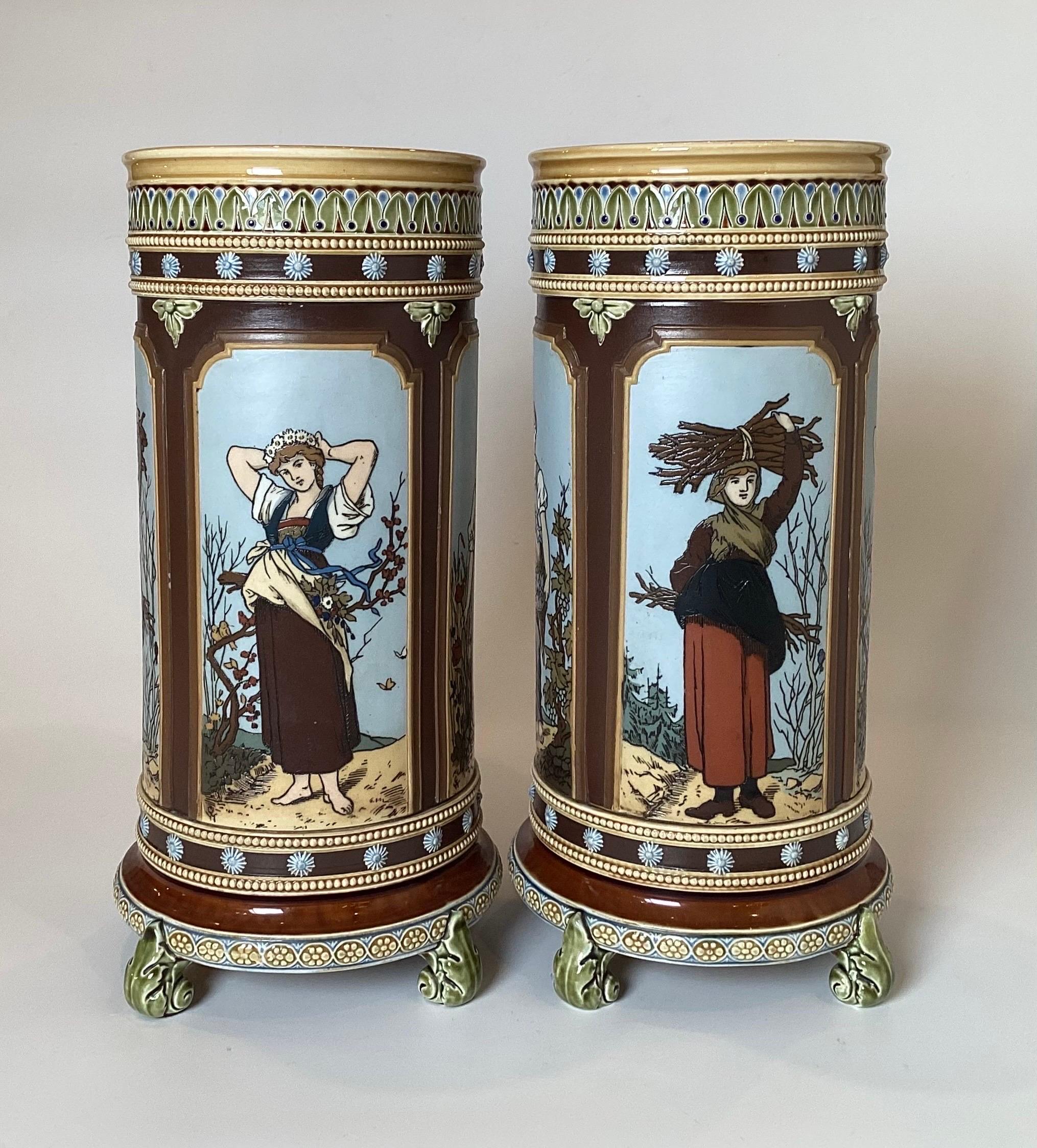 A pair of hand painted Mettlach footed cylinder vases, one depicting a maiden collecting tinder, the other a maiden gathering grapes for wine making. In overall very good condition. The colorful vases made in Germany, 1880s.
