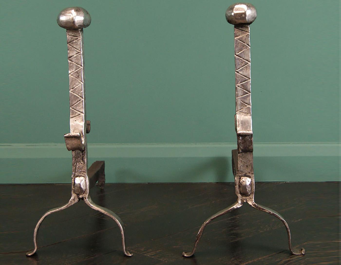 A pair of early nineteenth century polished wrought-iron fireplace andirons with incising to front face of standard, front and rear facing spit-hooks and segmented bulbous finials uppermost. All supported on arched supports. The riveted rear
