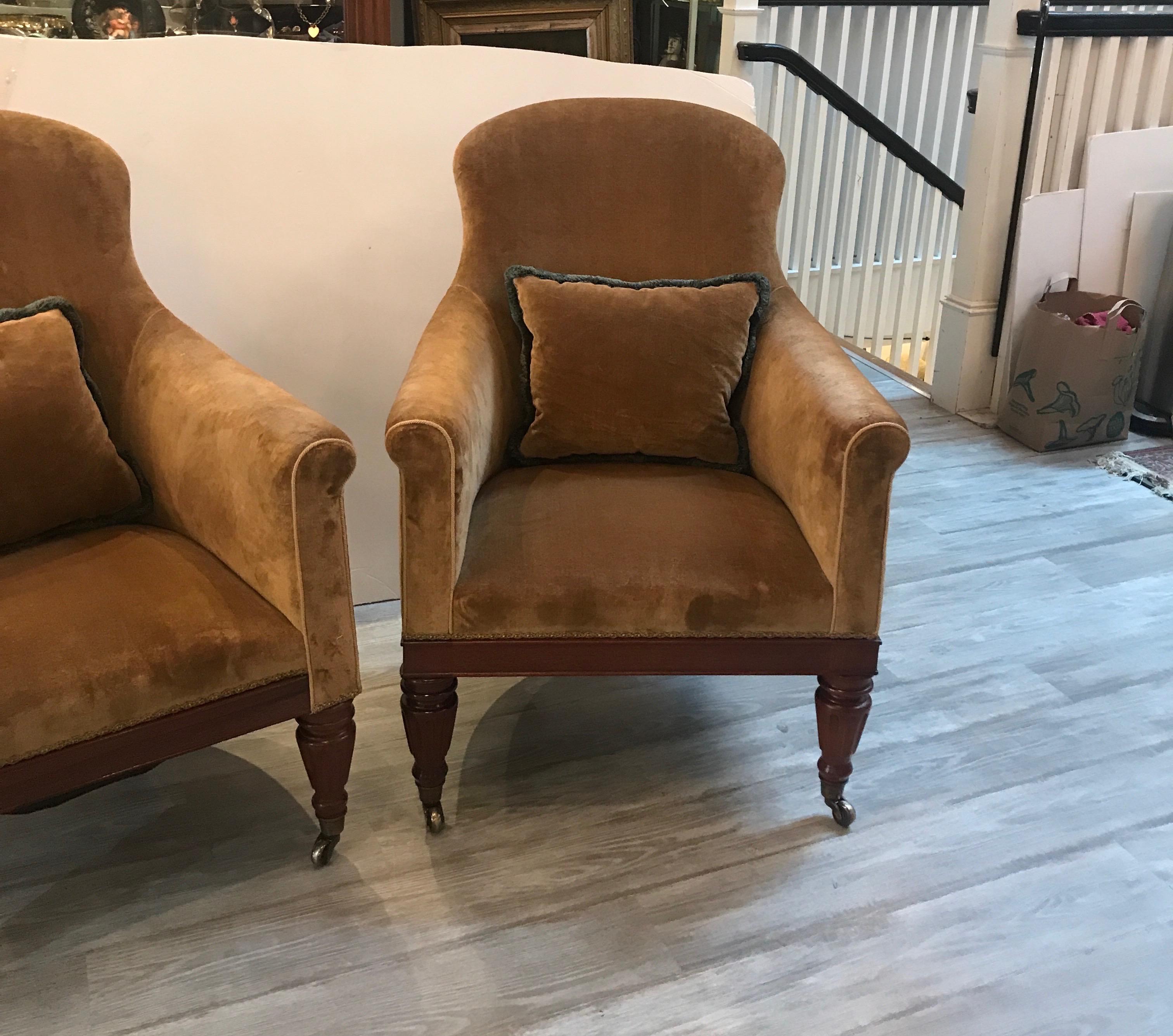 A pair of high back tub lounge chairs with tobacco colored velvet fabric. The mahogany base with original brass castors. The fabric which has some age in good condition but the chairs would look excellent with reupholstering, circa 1920.