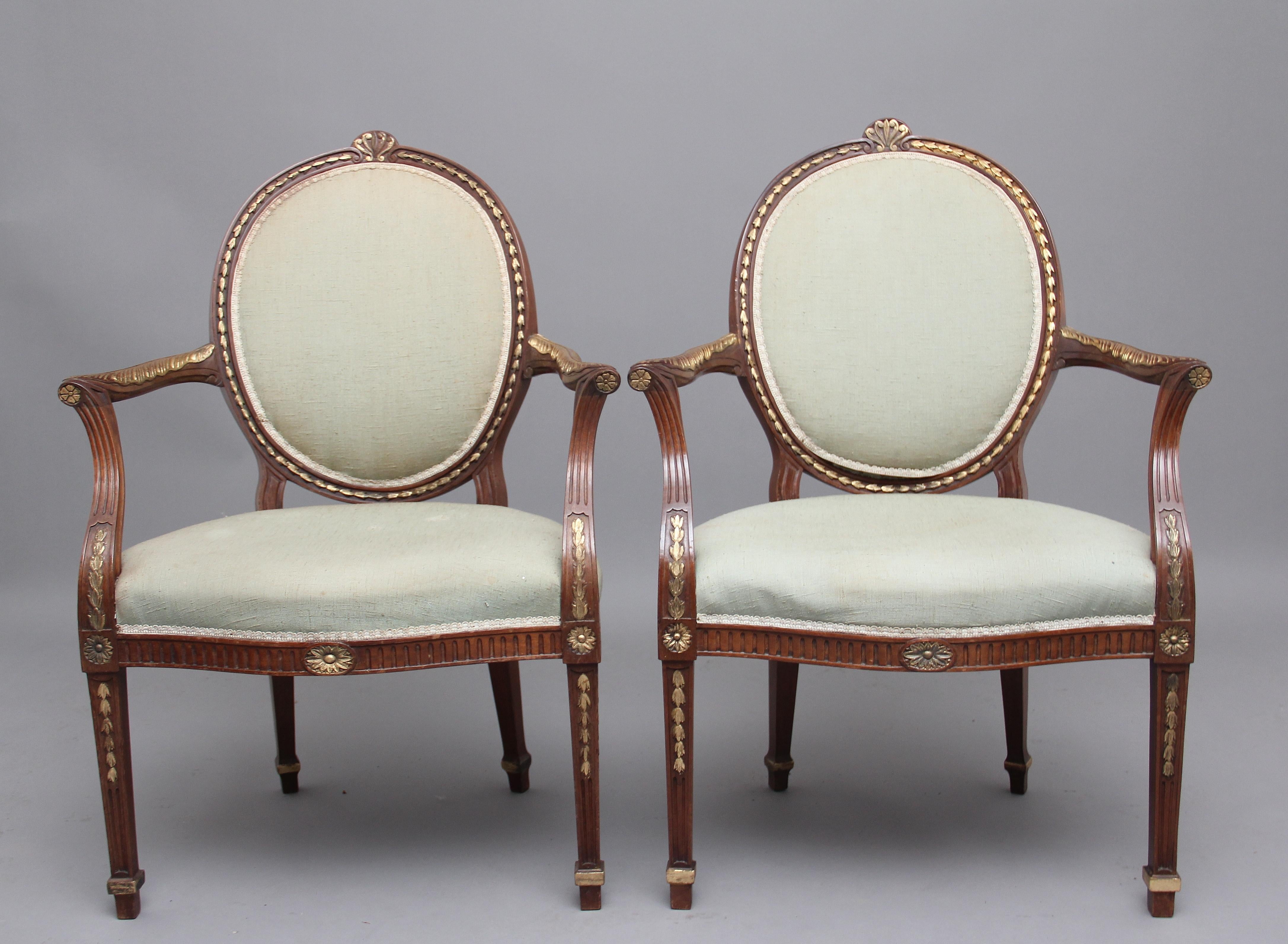 A decorative pair of early 20th century mahogany open armchairs in the Hepplewhite style, profusely decorated all over with gilding on carved wood, the oval padded backs and upholstered seat in a grey fabric, arms supported on out swept uprights,