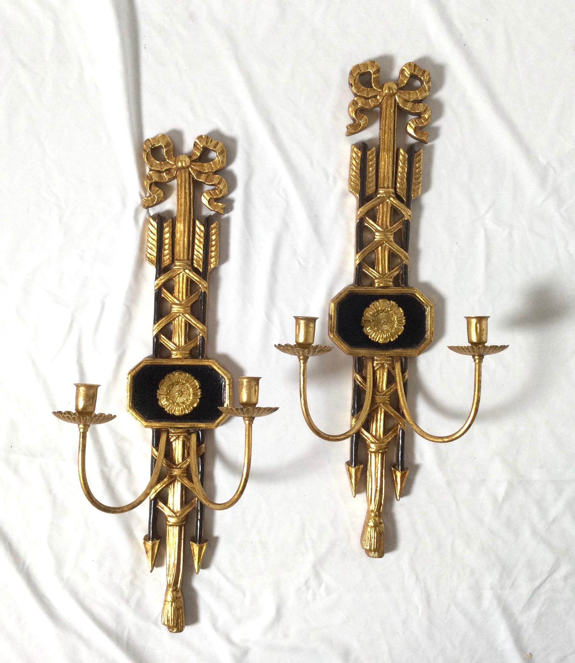 A pair of neoclassical styled Italian gilt and ebonized wood sconces with brass arms for two candles. The back plate is carved wood with bow tops with arrow motif... These are not electrified and for use with candles.