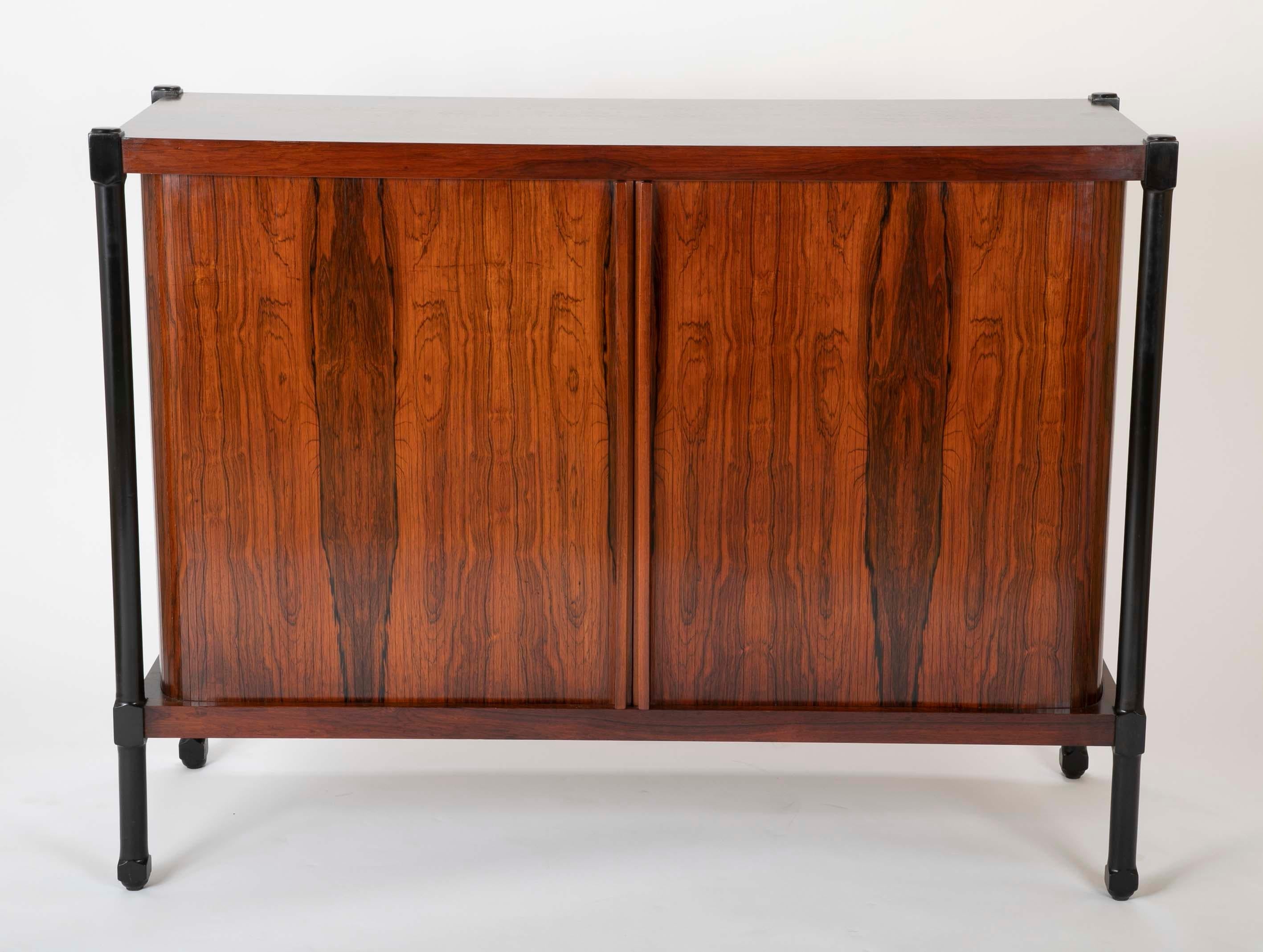 A striking pair of rosewood cabinets with tambour doors rising on ebonized legs. One cabinet with six drawers and two shelves. The other cabinet has four shelves. Unmarked.