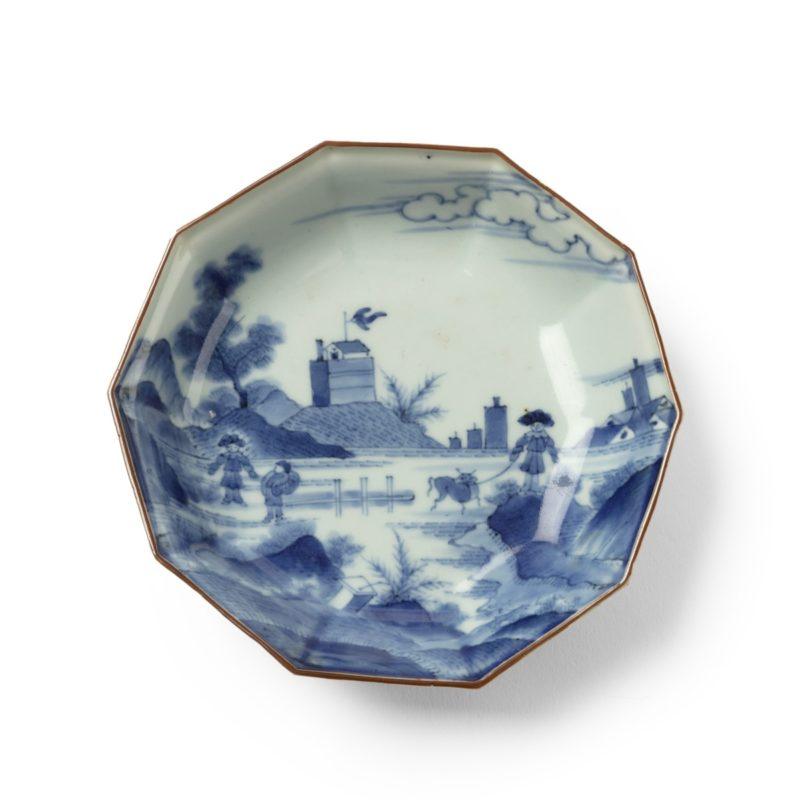 Each of these decagonal saucer-dishes is painted in underglaze-blue with a landscape of two Dutchmen and a boy warmly wrapped up and wearing large hats, two appear to be skating and one has a cow on a rope, in front of a hill fort flying a flag and