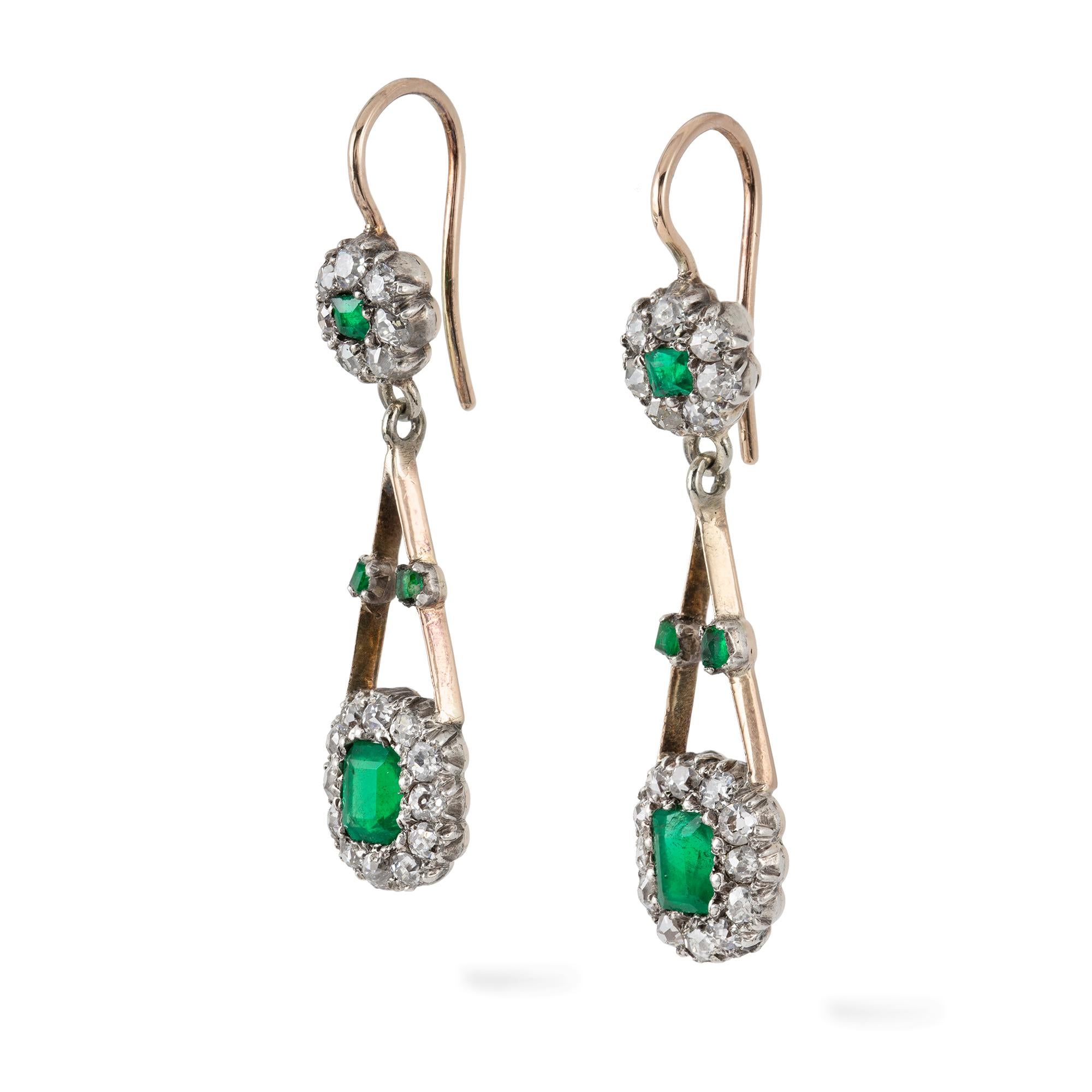 A pair of Edwardian emerald and diamond earrings, each earring with a cushion-cut emerald surrounded by twelve old-cut diamonds attached to two knife-edged gold bars forming a V, suspended by a cluster consisted of a square-cut emerald and seven