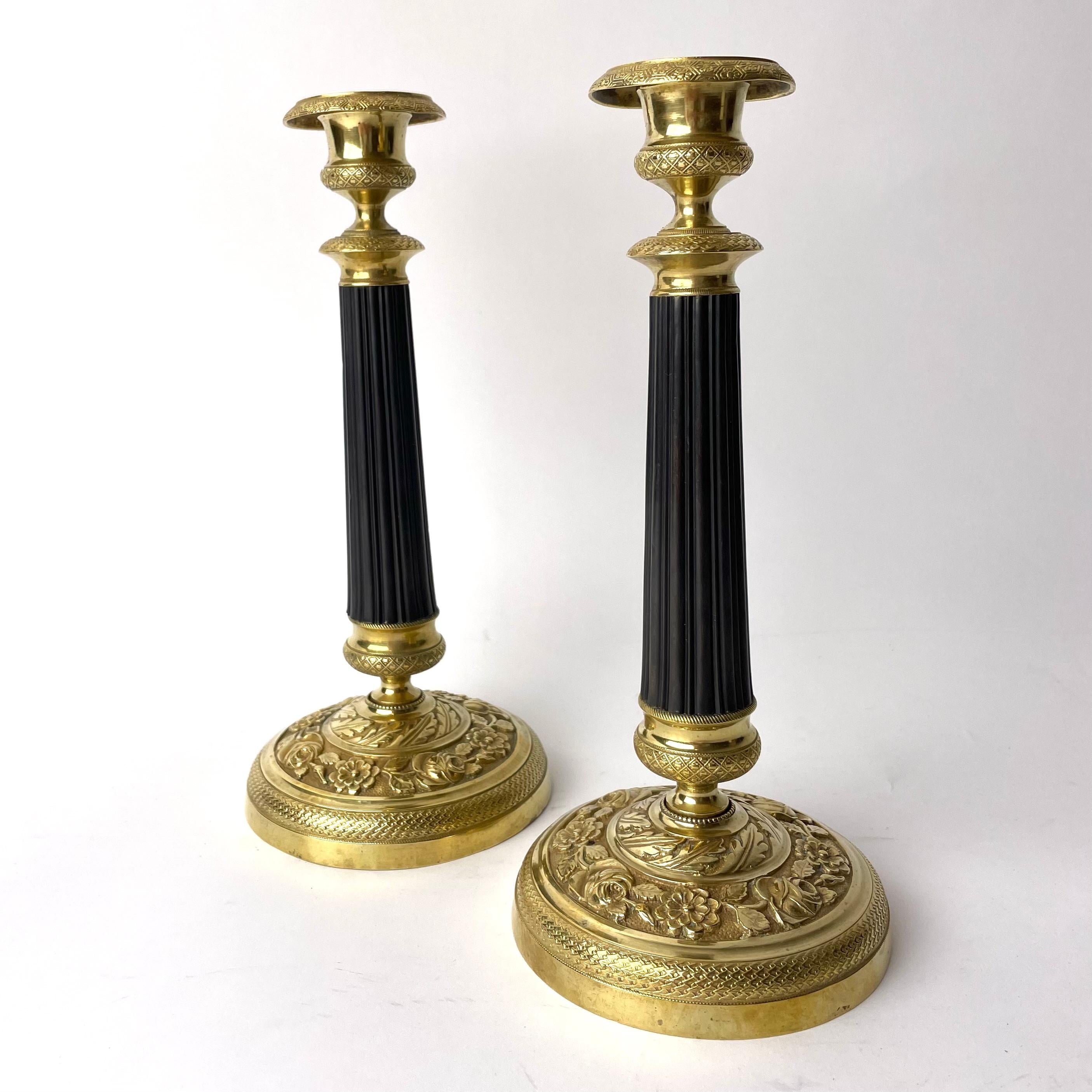 A pair of exquisite Empire candlesticks which combine gilt and patinated brass. Made in 1820s France. Various intricate rose, floral and leaf patterns adorn the bases in gilt brass. Emblematic of the Empire period, these candlesticks feature fluted