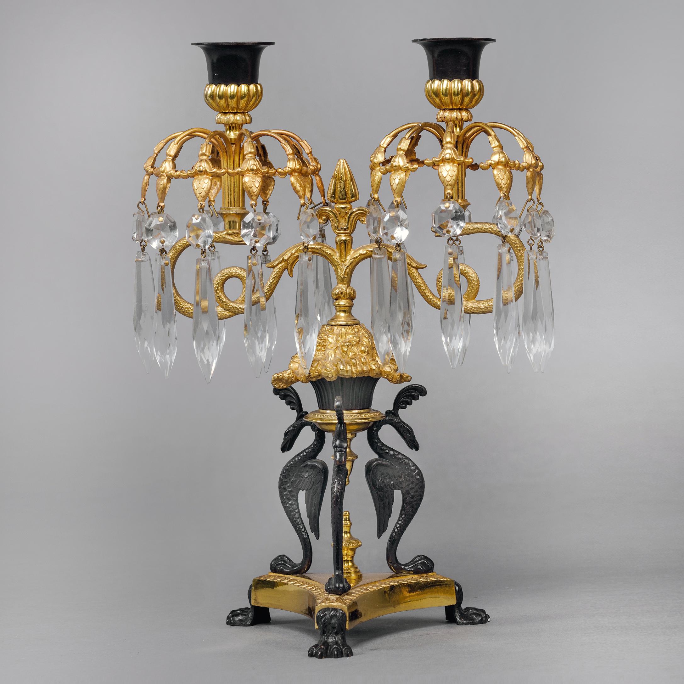 A pair of Empire revival gilt and patinated bronze twin-light lustre candelabra.

Each candelabra has a concave tripartite base with winged paw feet, supporting three gilt-bronze eagle headed chimera, below grape and vine leaf cast socles with