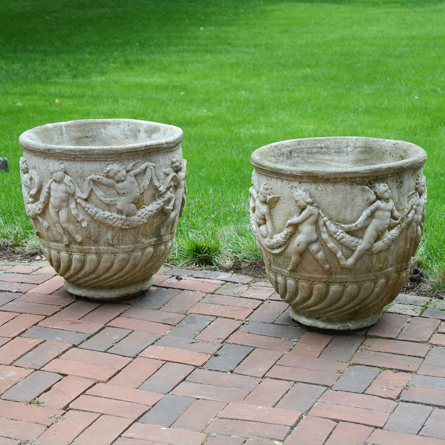 A pair of composition stone planters ornamented with frolicking cherubs, and swags, both ribbon and floral, English, circa 1960. Measures: 21 ins. high, 21 ins. diameter, 12 ins. diameter at base.