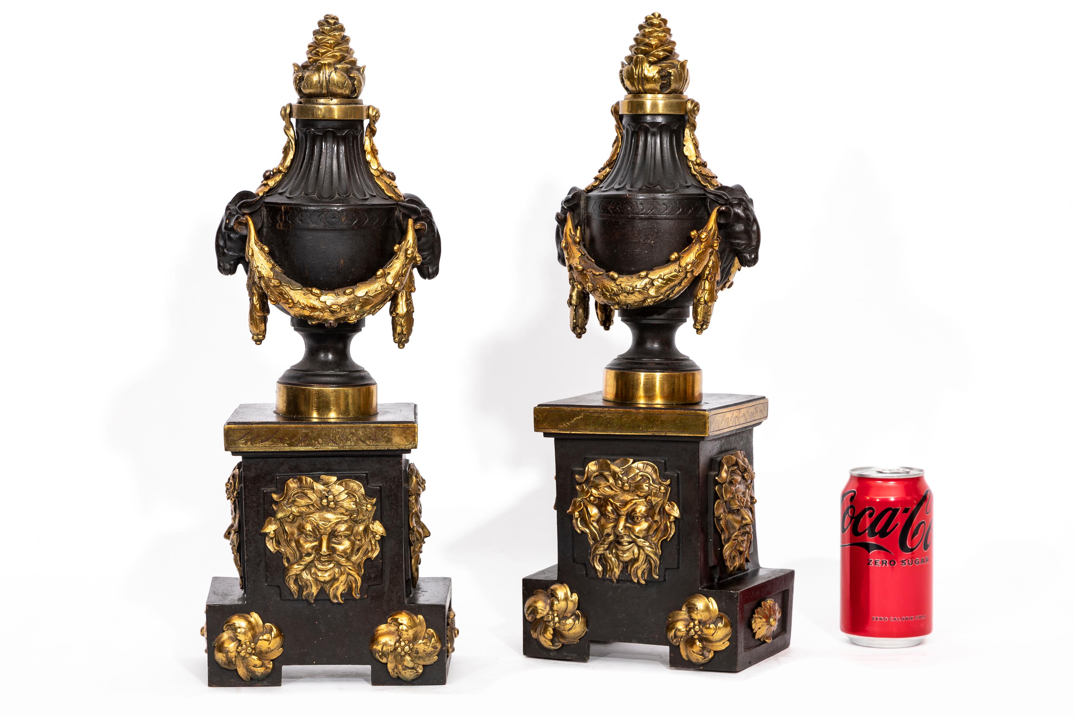 A Pair of English Regency Period Patinated and Ormolu Rams Head Andirons.  Each is beautifully patinated with a dark patina and further accented with ormolu fittings.  The body is of urn form with ormolu acorn finial tops, ormolu wreaths and