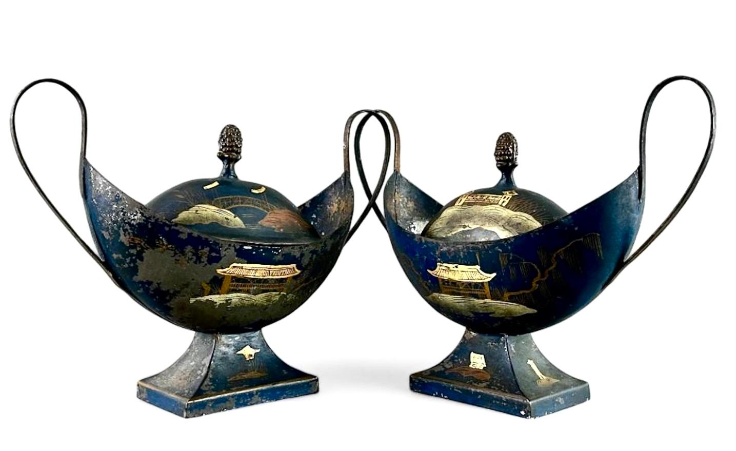 A Pair of 19th century English Regency Tole Peinte Chestnut Urns. Urns feature ovoid forms with delicate scroll arms and domed covers with pine cone finial. Urns are painted with japanned decoration in navy blue and gilding. Can be used as